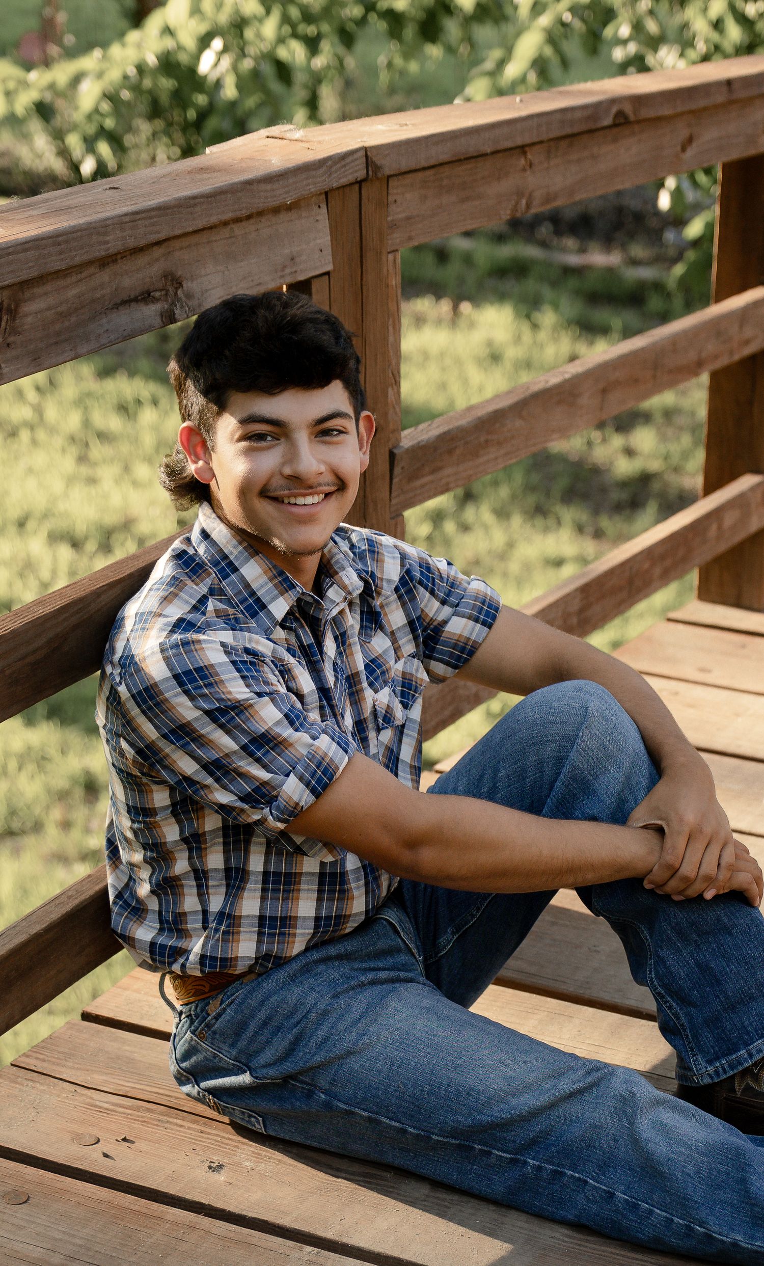 Smiling teenage boy in plaid shirt and jeans sitting on wooden bridge steps outdoors.