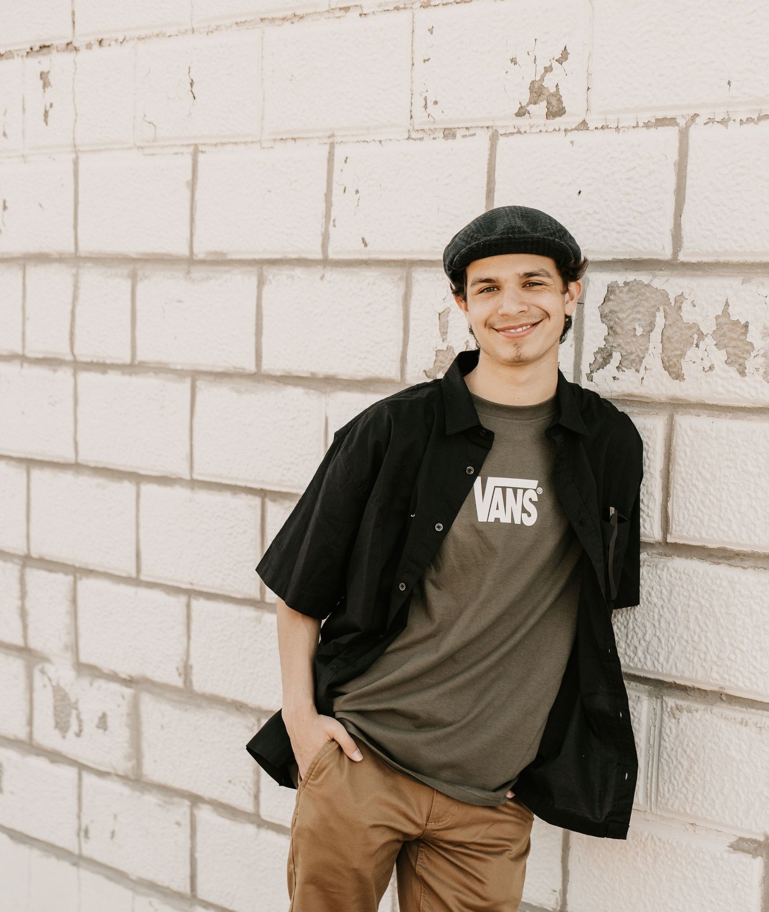 Young man smiling in Vans t-shirt, black cap and khaki pants, leaning against white brick wall.