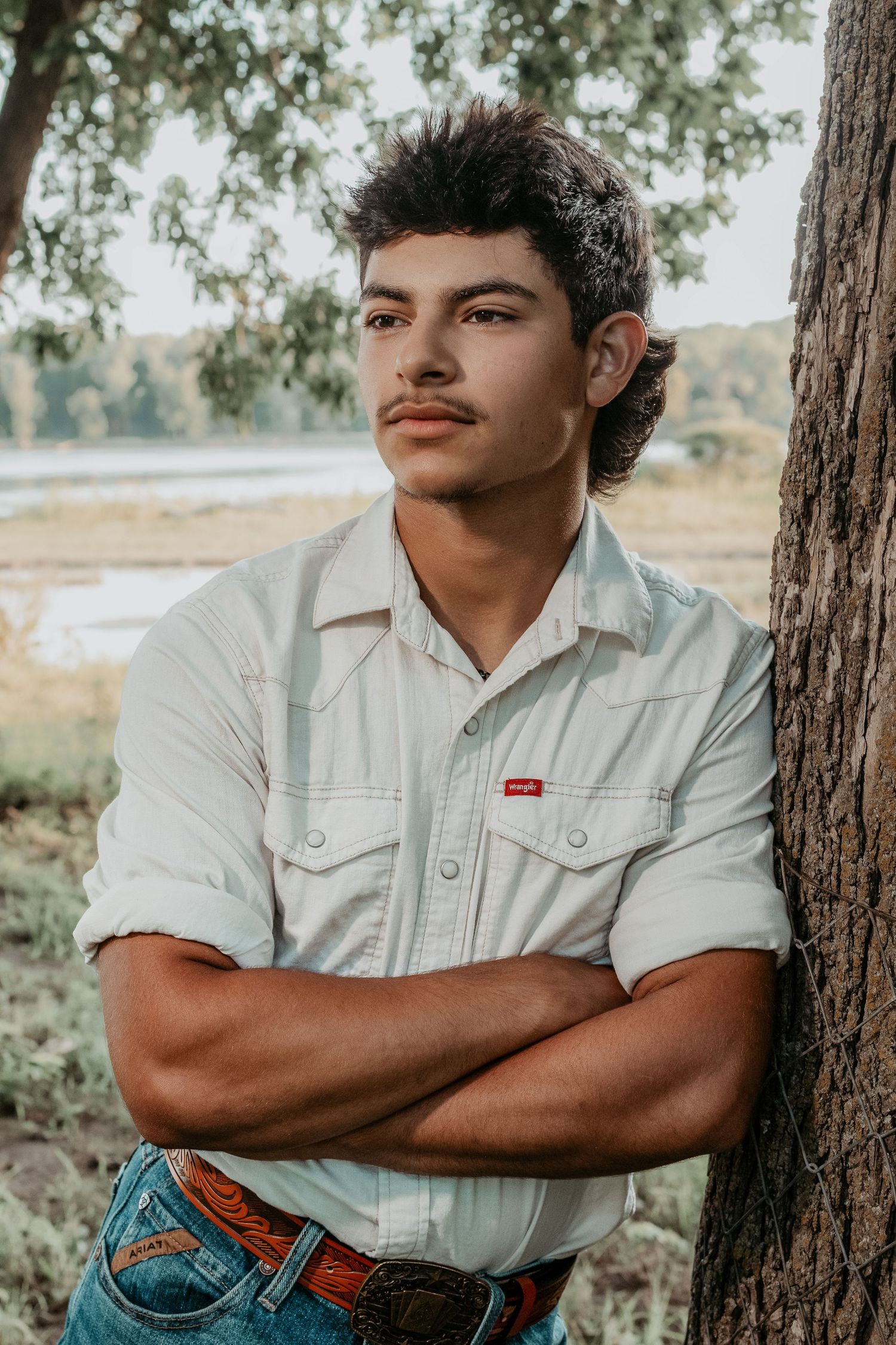 A young man in a Levi's shirt stands with arms crossed leaning against a tree in an outdoor setting.