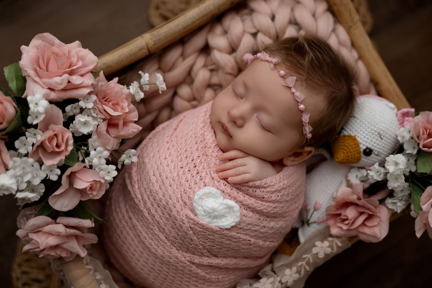Newborn baby swaddled in pink wrap sleeping in a wicker basket surrounded by pink roses and floral headband.