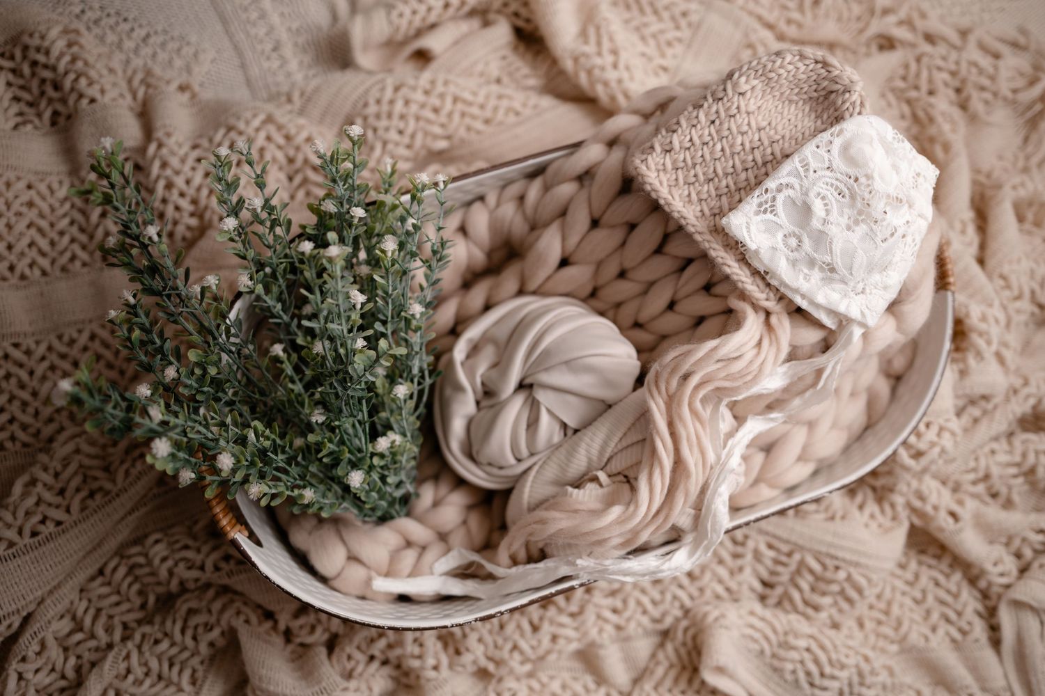 Delicate newborn photography setup with soft beige wraps, knit blanket and fresh greenery on white bowl.