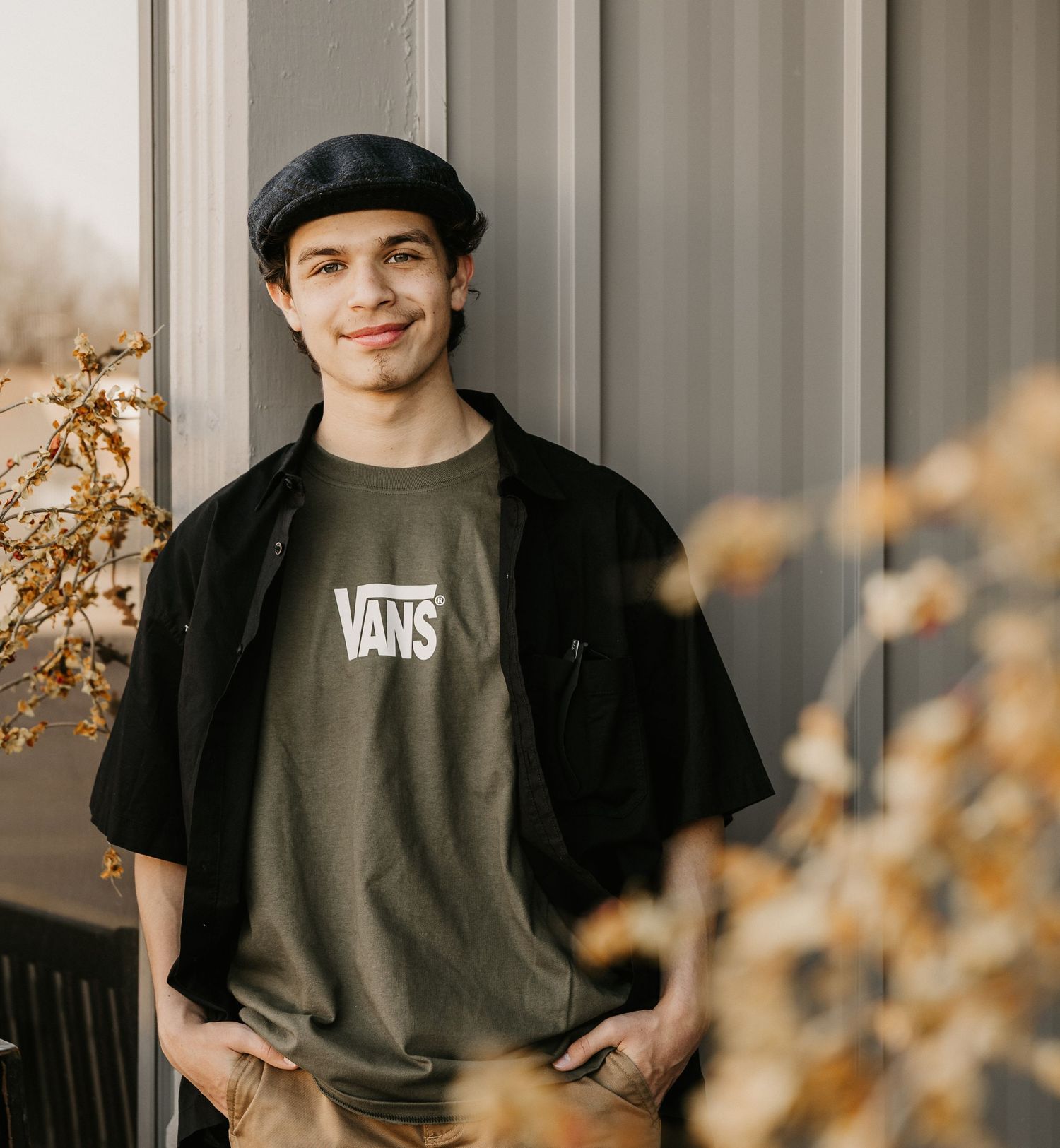 Young man in Vans t-shirt and black cap smiling outdoors near autumn foliage and gray wall.