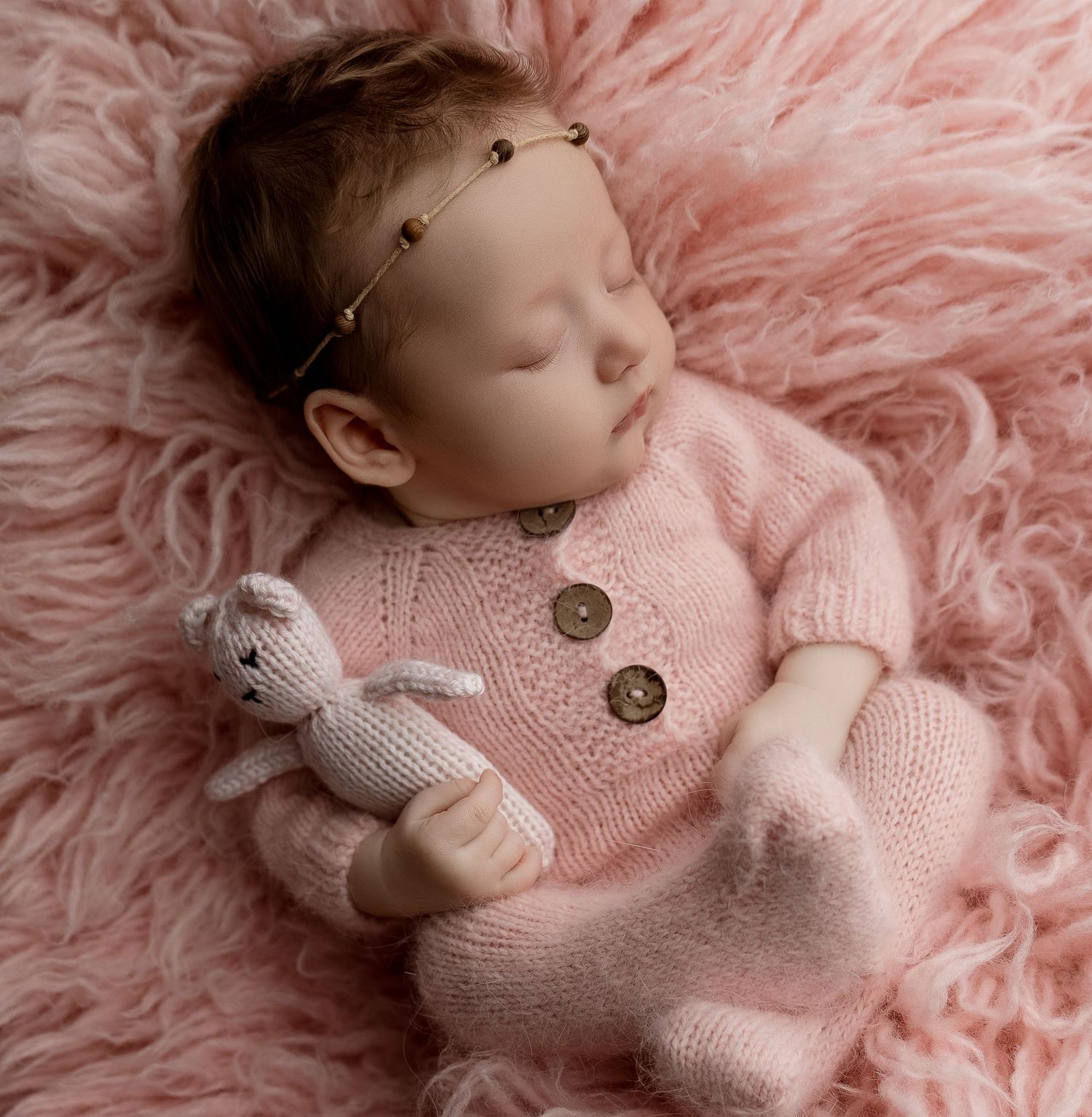 Sleeping newborn baby in pink knit outfit holding a bunny toy on a fluffy pink backdrop.