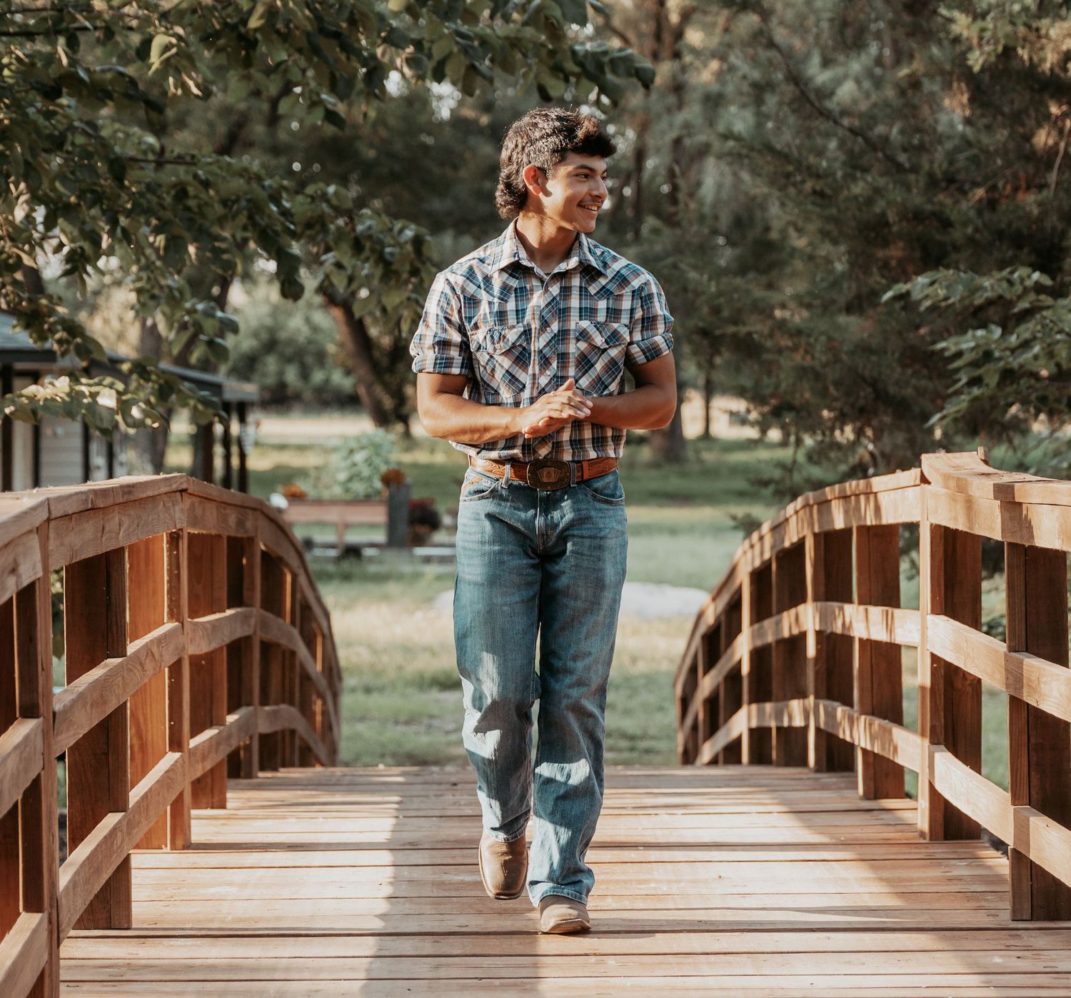 Man in plaid shirt and jeans walking on a wooden arched bridge surrounded by lush green trees.