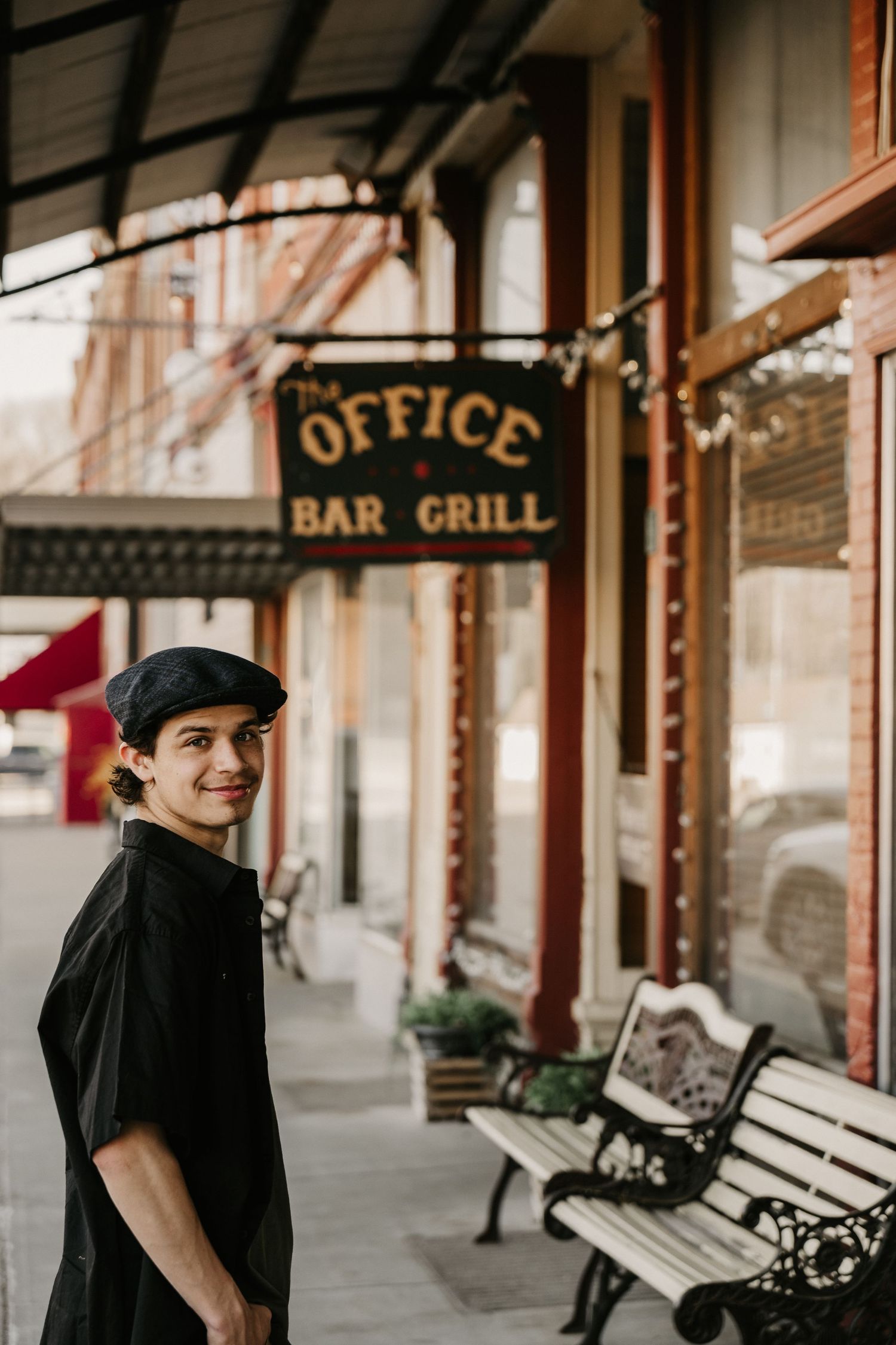 A smiling person in black outfit and beret stands outside The Office Bar Grill with benches on a charming small-town sidewalk.