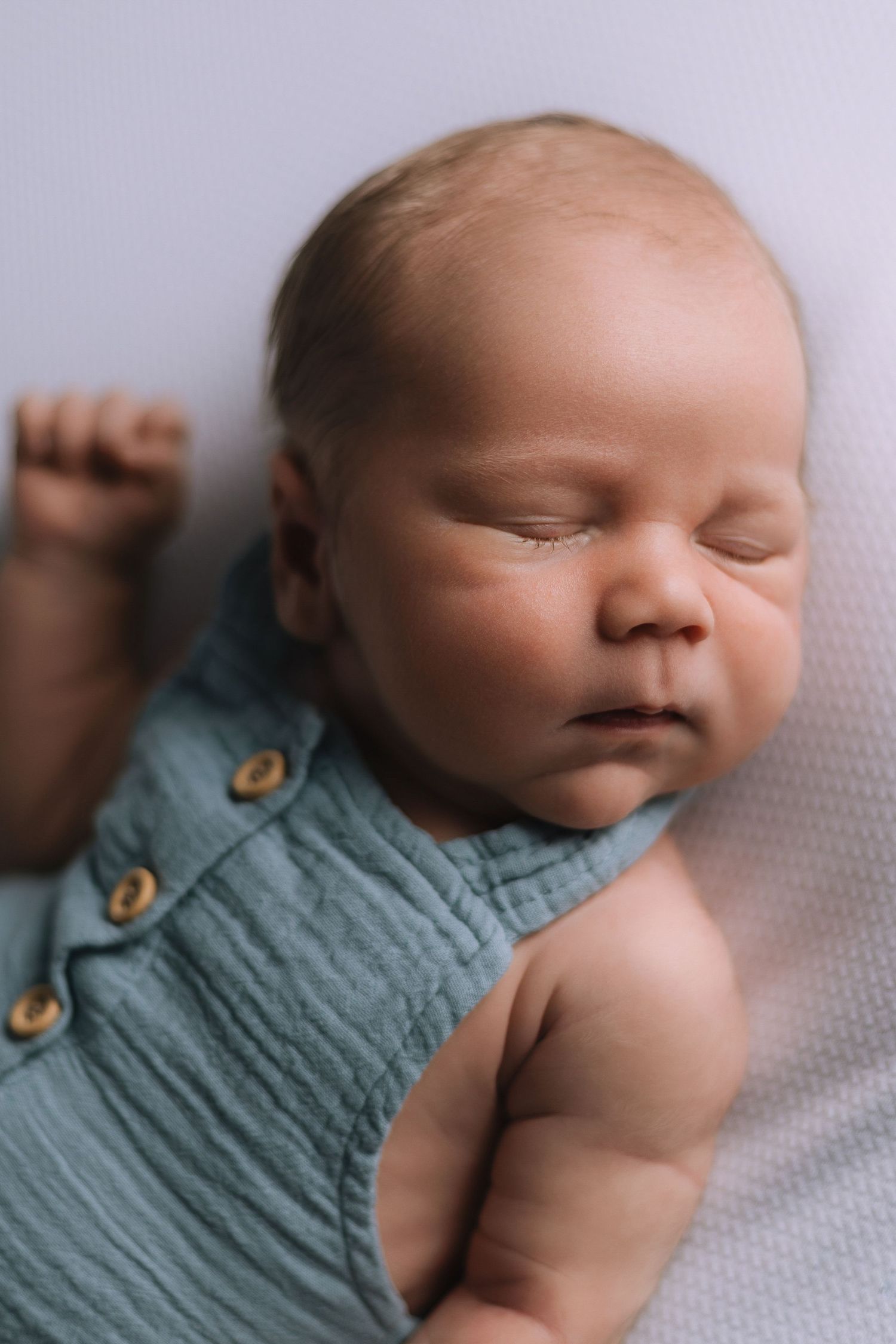 Sleeping newborn baby in teal muslin romper lying on white blanket, peaceful infant portrait photography.