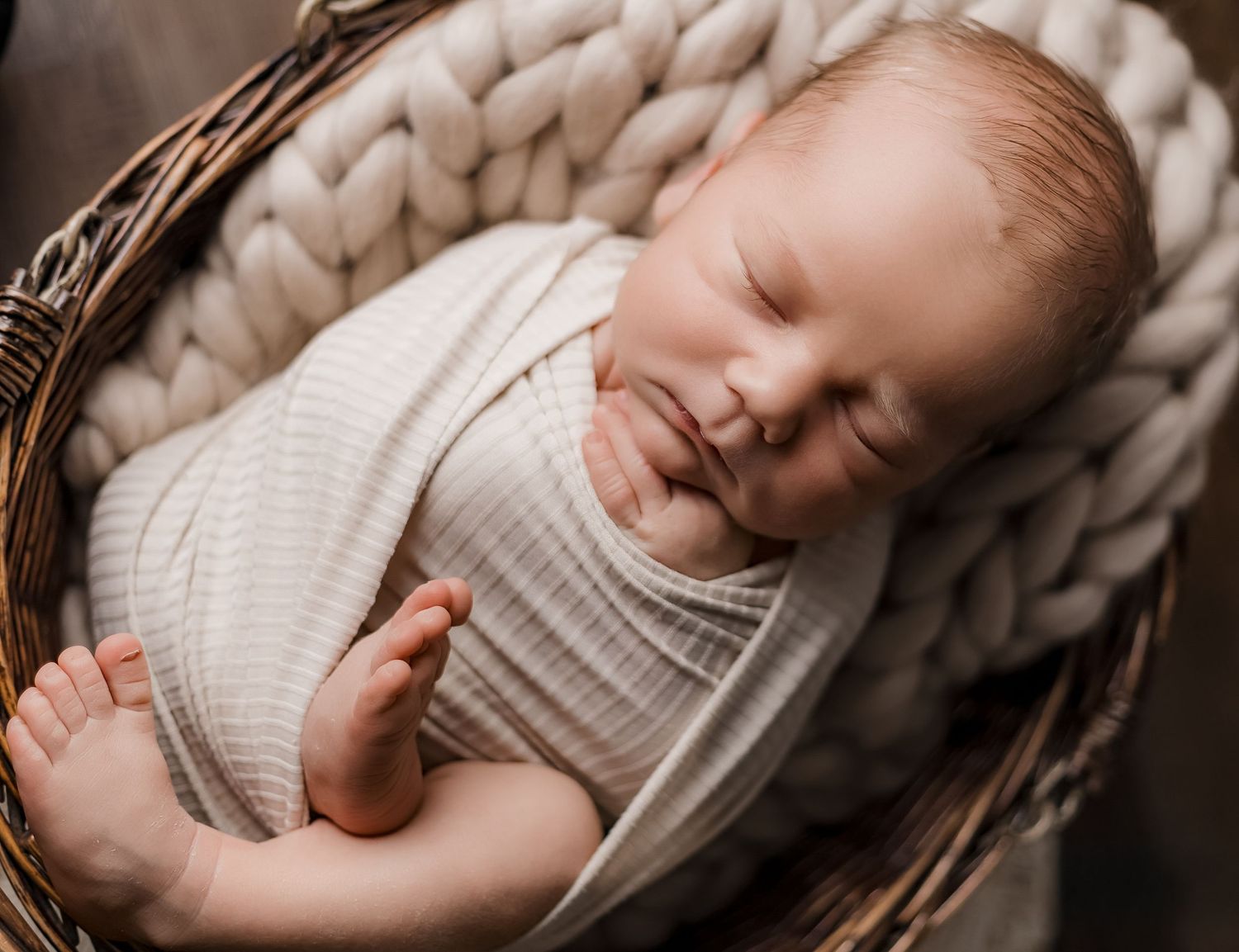 Peaceful newborn baby swaddled in white wrap sleeping in a rustic wicker basket with chunky knit blanket.