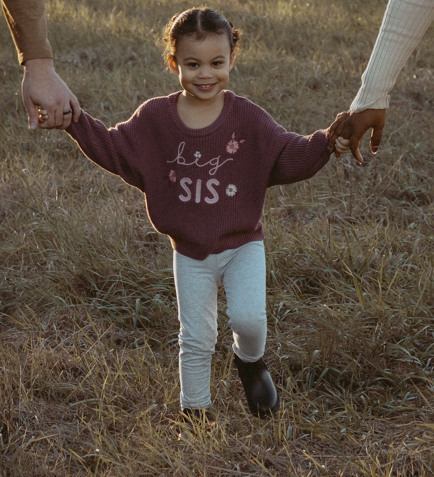 A happy girl in a Big Sis sweatshirt walks holding hands with parents in a sunny field.
