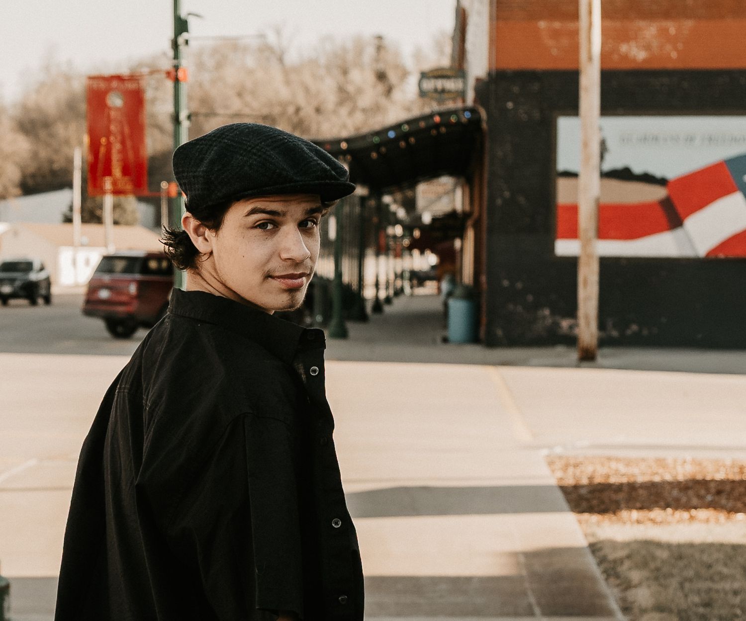 Young man in black coat and beret hat standing on urban street sidewalk, looking back over shoulder.