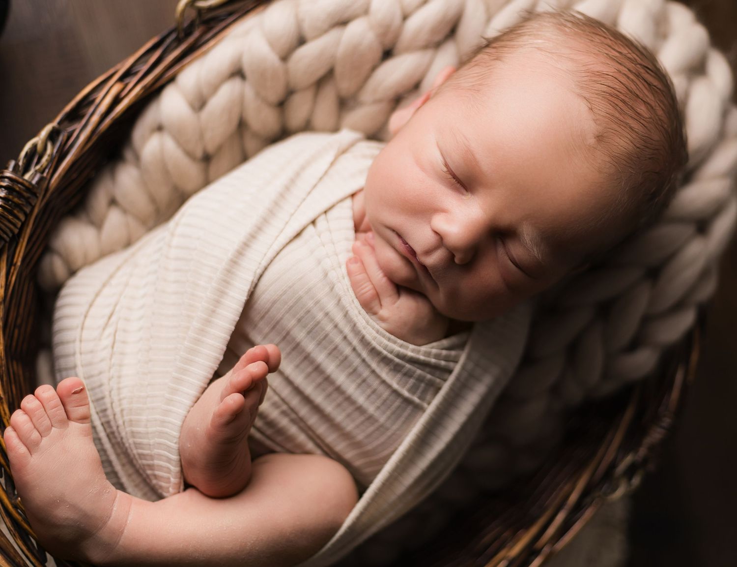 Peaceful newborn baby swaddled in cream wrap sleeping in a rustic wicker basket with chunky knit blanket.