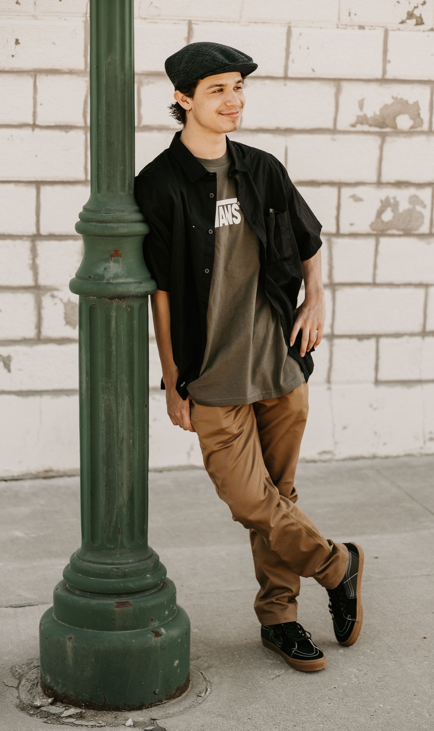 Young man in black jacket, beret, Vans tee, tan pants, leaning on green lamp post against brick wall.