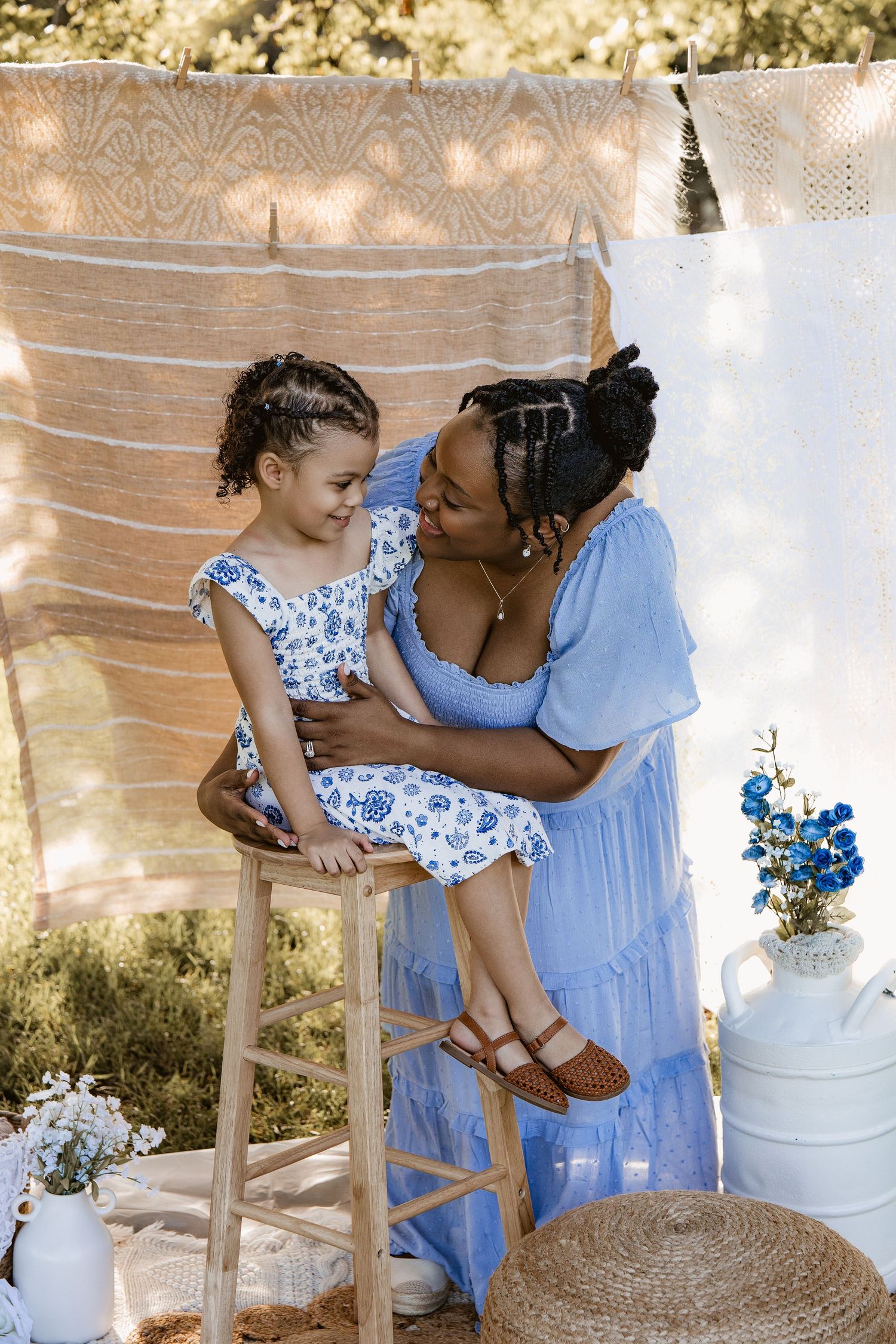 A mother in a blue dress holds her young daughter on a stool outdoors, both wearing matching blue floral outfits.