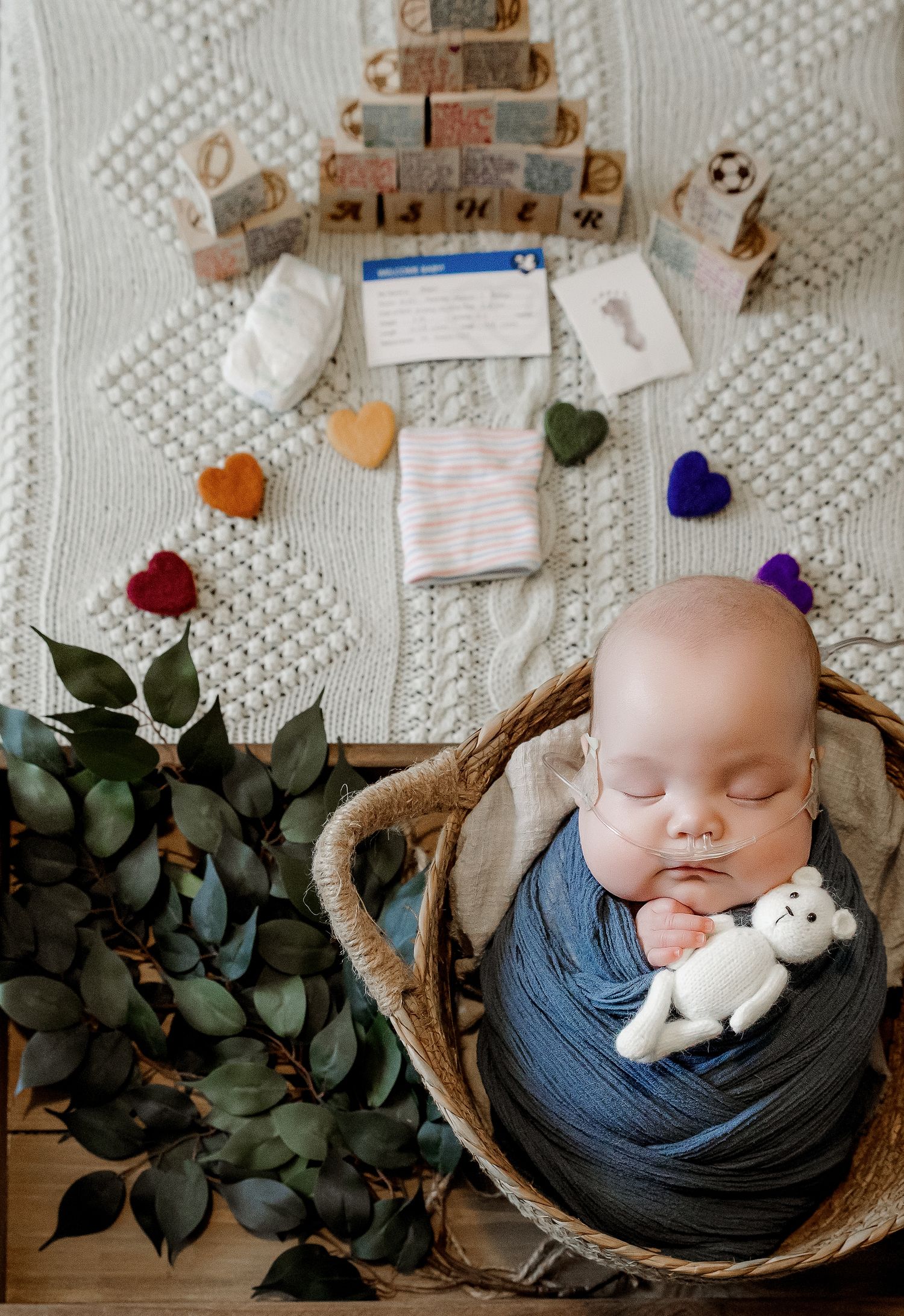 Sleeping newborn baby wrapped in blue swaddle in wicker basket with toy bear, surrounded by blocks and colorful felt hearts.