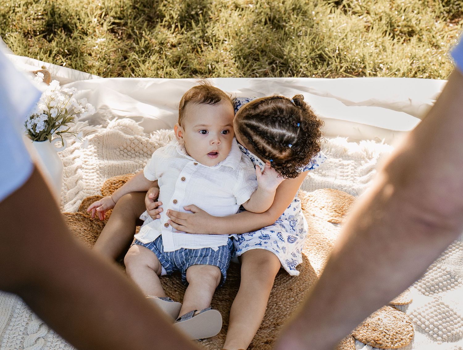 A toddler girl lovingly kisses her baby brother while they sit together on a cozy outdoor blanket.
