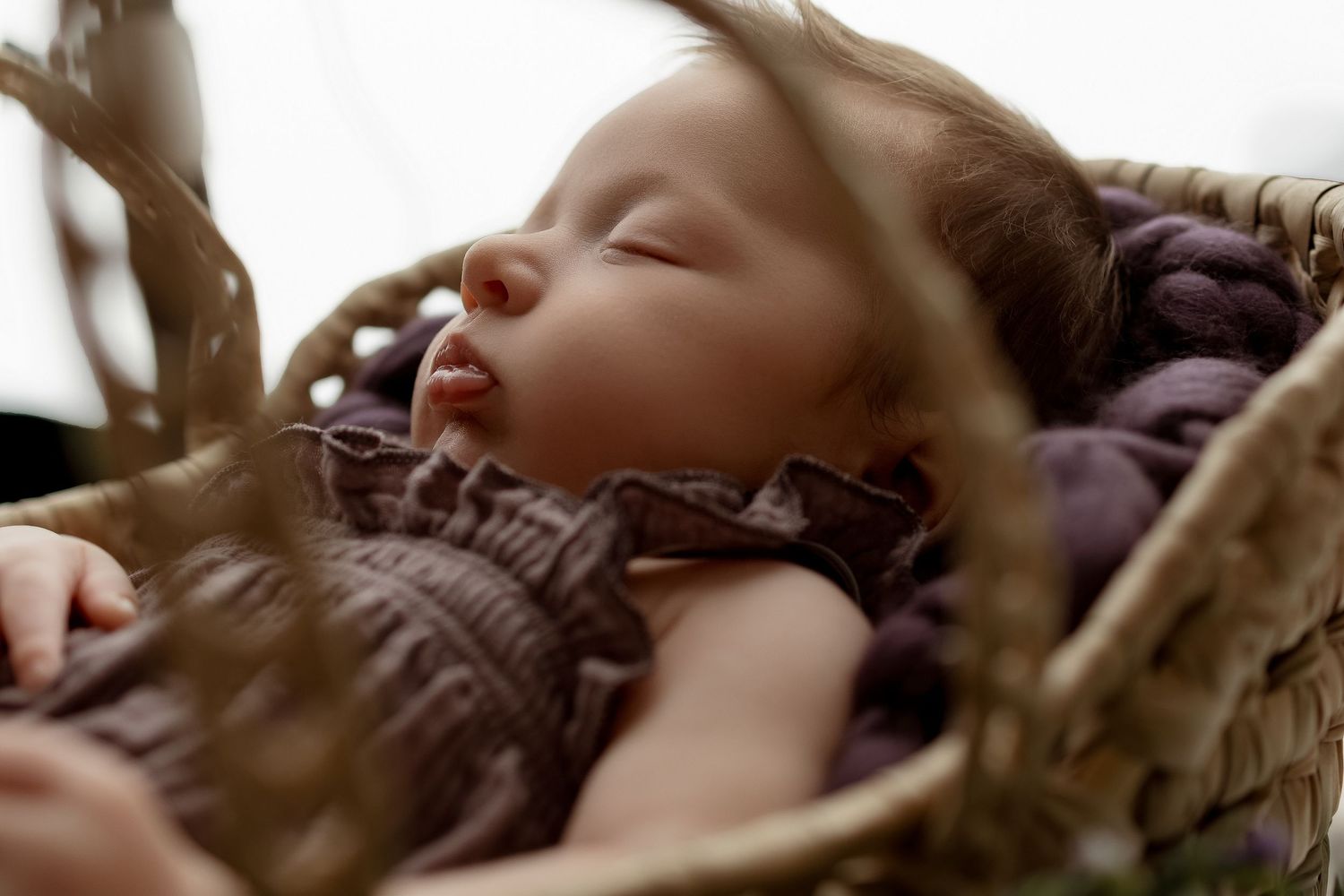 Sleeping newborn baby nestled in purple fabric inside a rustic wicker basket.