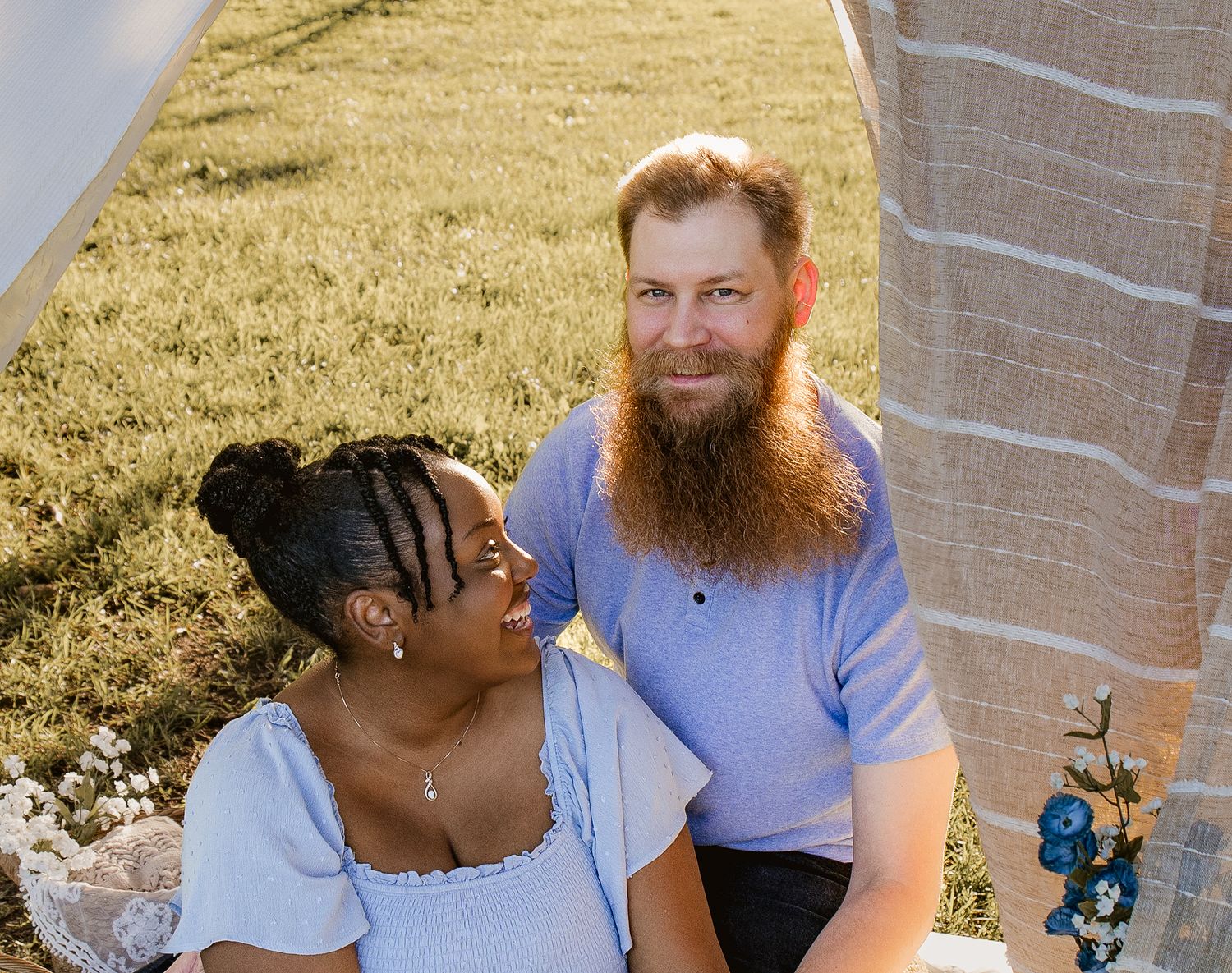 A smiling interracial couple sits outdoors on grass, the woman in a blue dress and the bearded man in a light blue shirt.