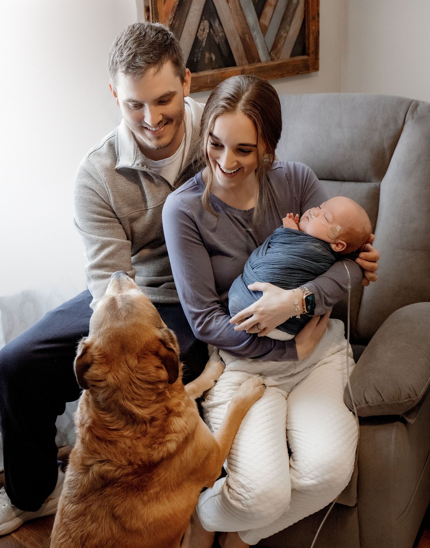 A smiling couple sits in a gray recliner with their newborn baby while a golden retriever dog looks up at them.