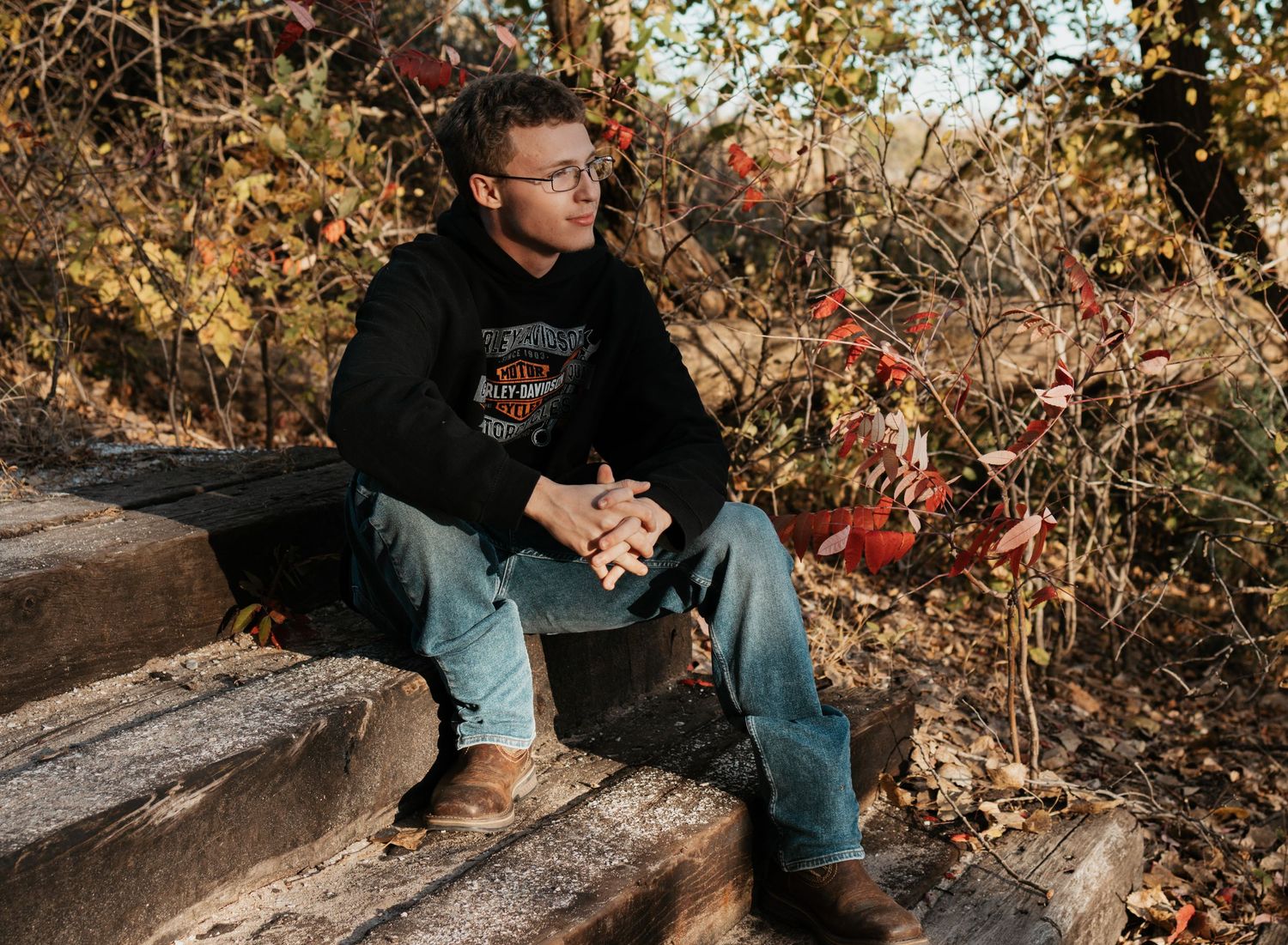 Young man in Harley-Davidson hoodie and jeans sitting on wooden steps outdoors among autumn foliage.