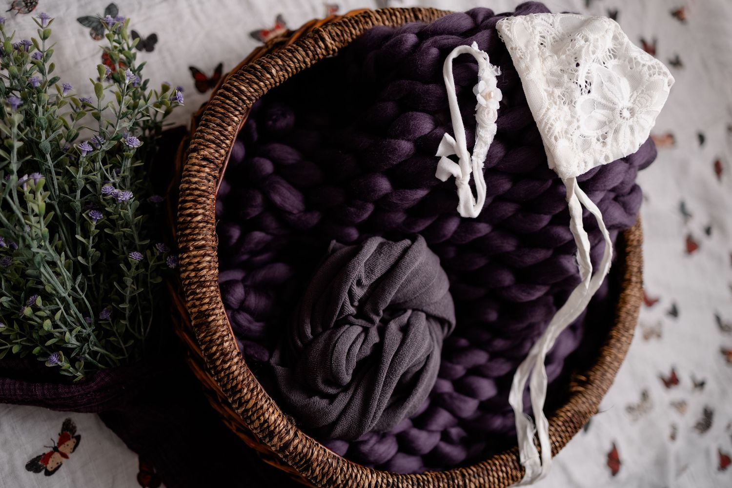 A woven basket filled with folded purple and gray fabric linens sits beside fresh herbs on a textured surface.