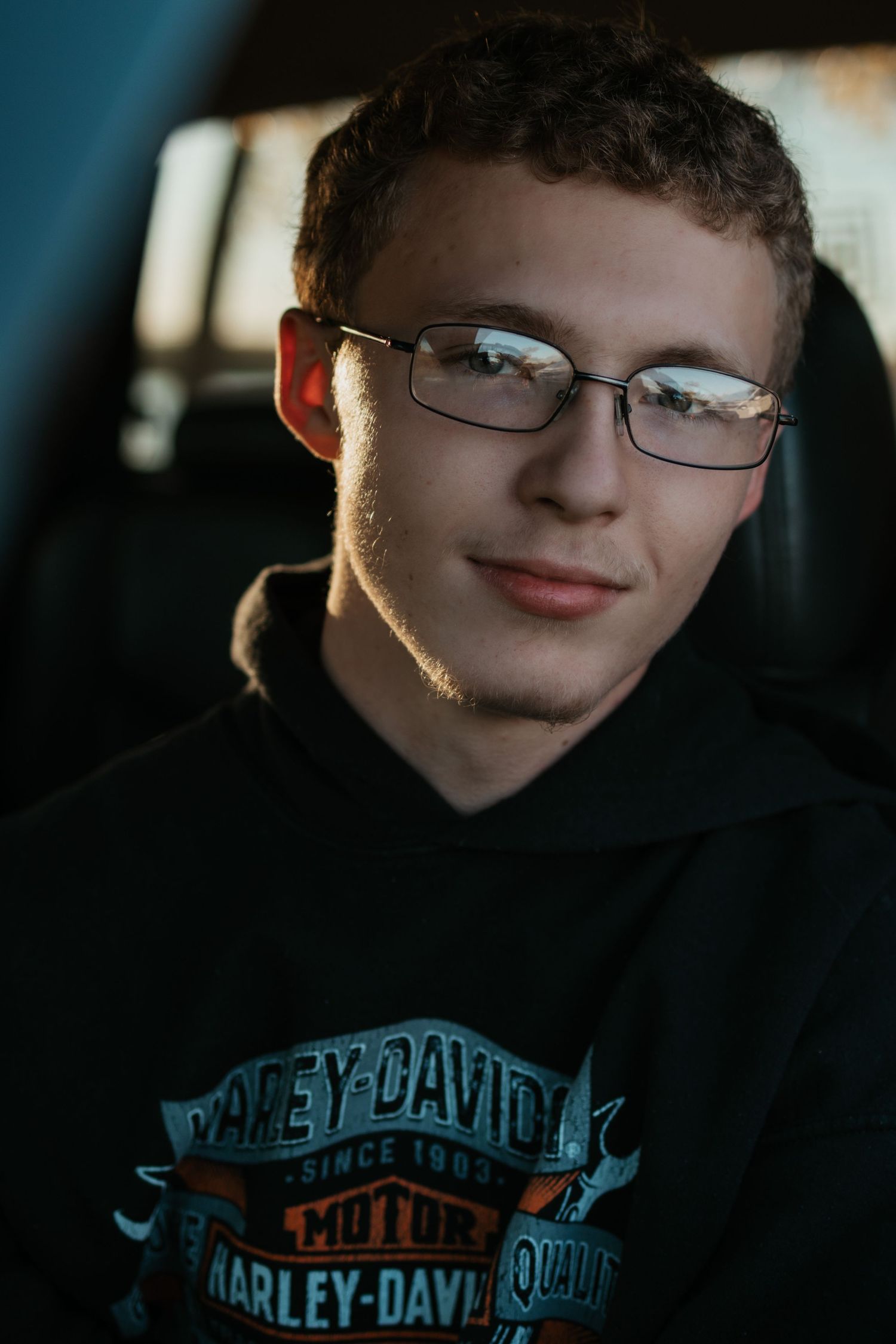 Young man wearing glasses and Harley-Davidson hoodie smiling inside a vehicle in warm light.