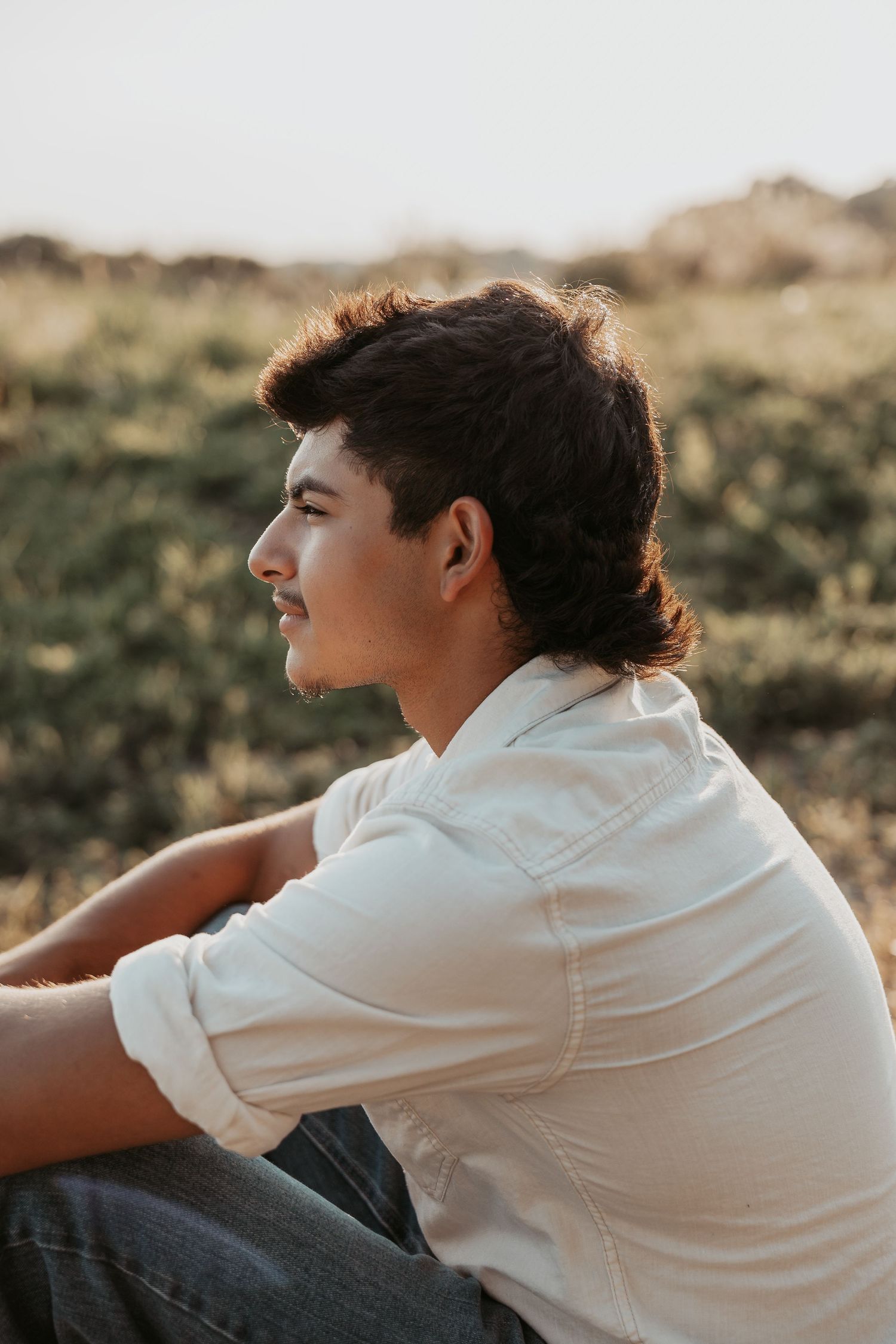 Young man in white shirt sitting outdoors in profile view, gazing thoughtfully into the distance at golden hour.