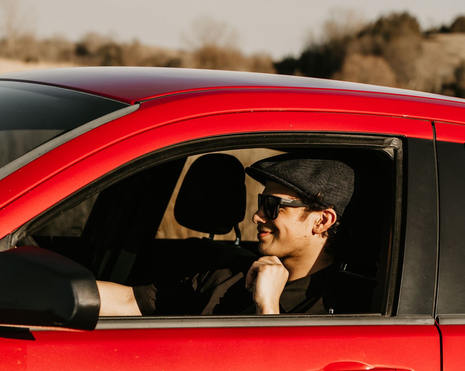 A stylish man wearing sunglasses and a flat cap sits in a red car, gazing thoughtfully out the window.