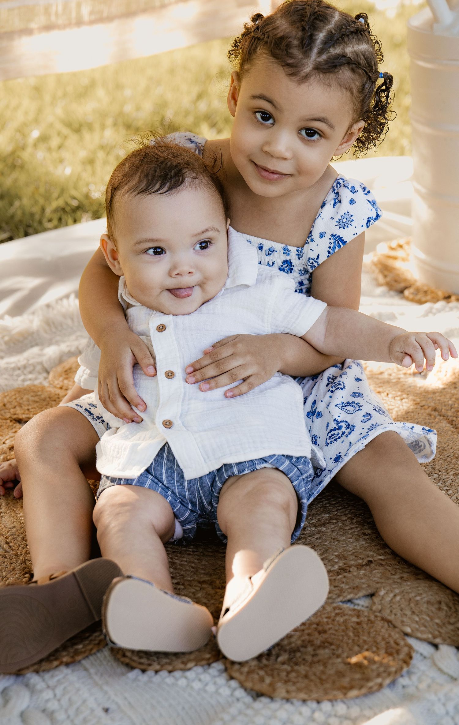 A toddler girl in a blue floral dress hugs a smiling baby boy in a white romper while sitting outdoors.