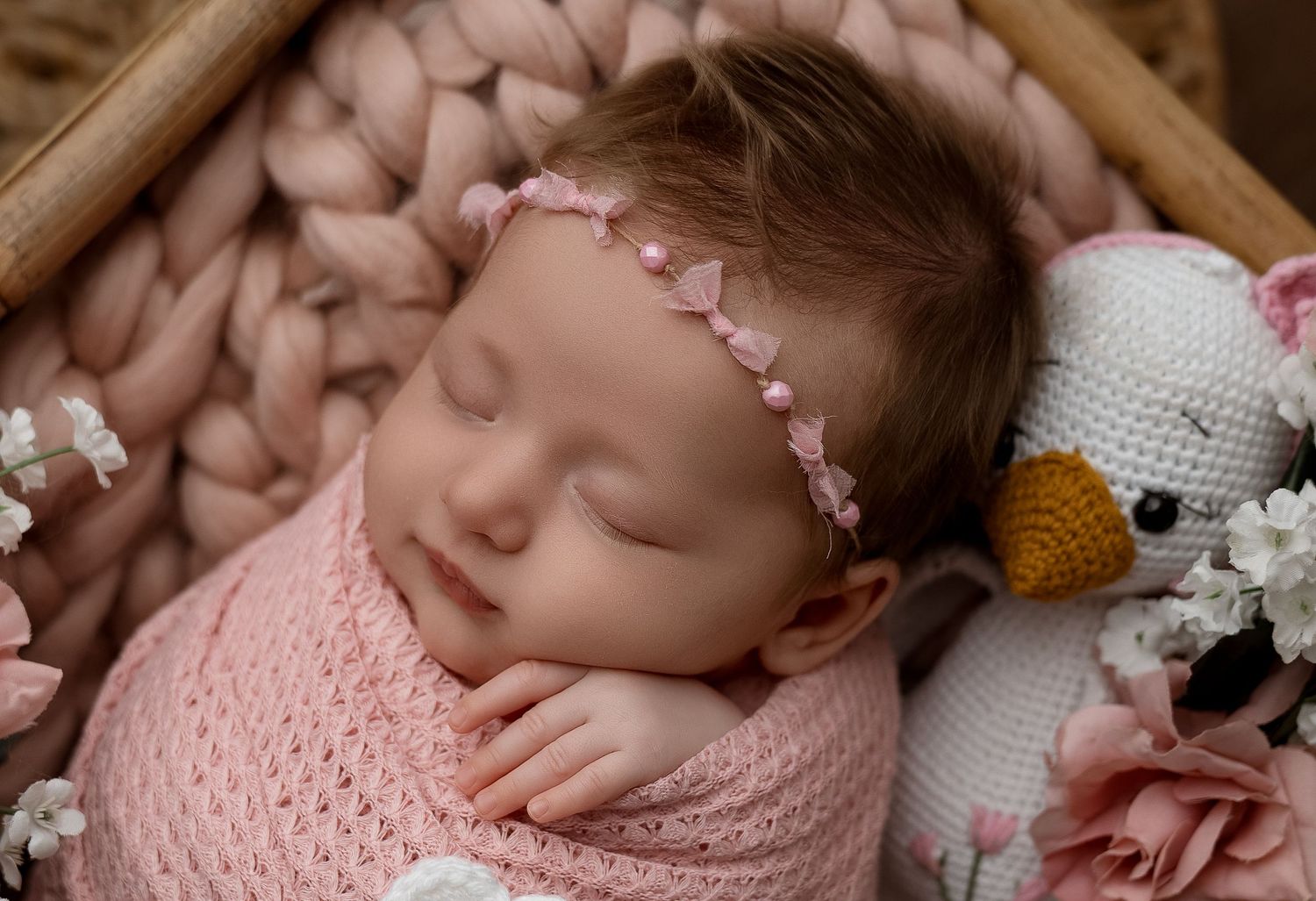 Sleeping newborn baby girl wrapped in pink knit swaddle with floral headband beside crochet bear toy.