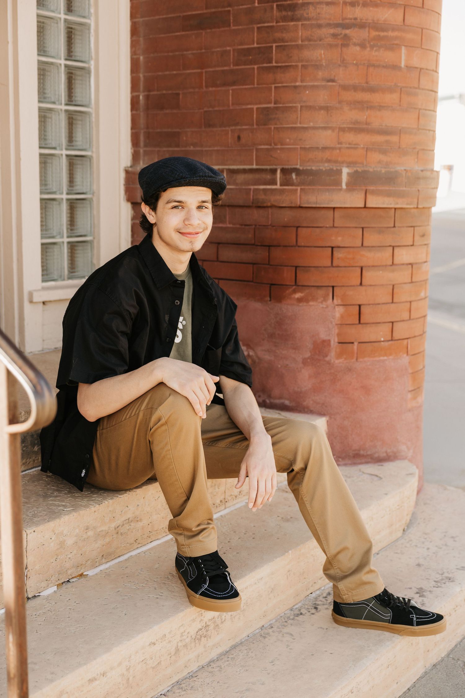Young man in black cap and jacket sits on brick building steps, smiling confidently in tan pants and dark sneakers.