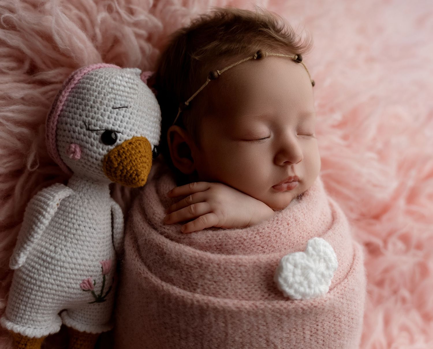 Sleeping newborn baby swaddled in pink wrap with crochet duck toy on fluffy pink background.