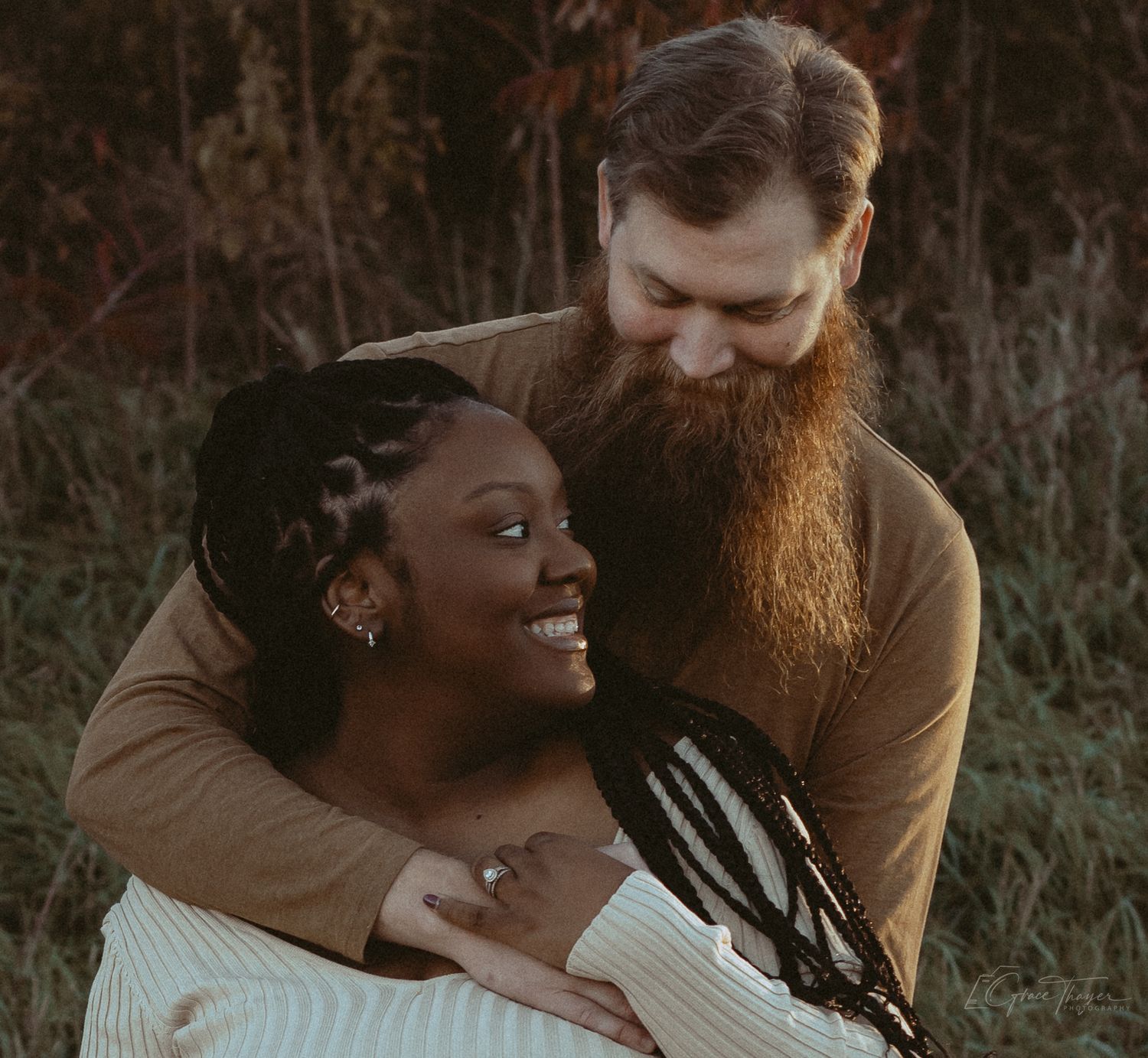 A smiling Black woman with braids embraces a bearded redhead man outdoors in a warm, romantic couples portrait.