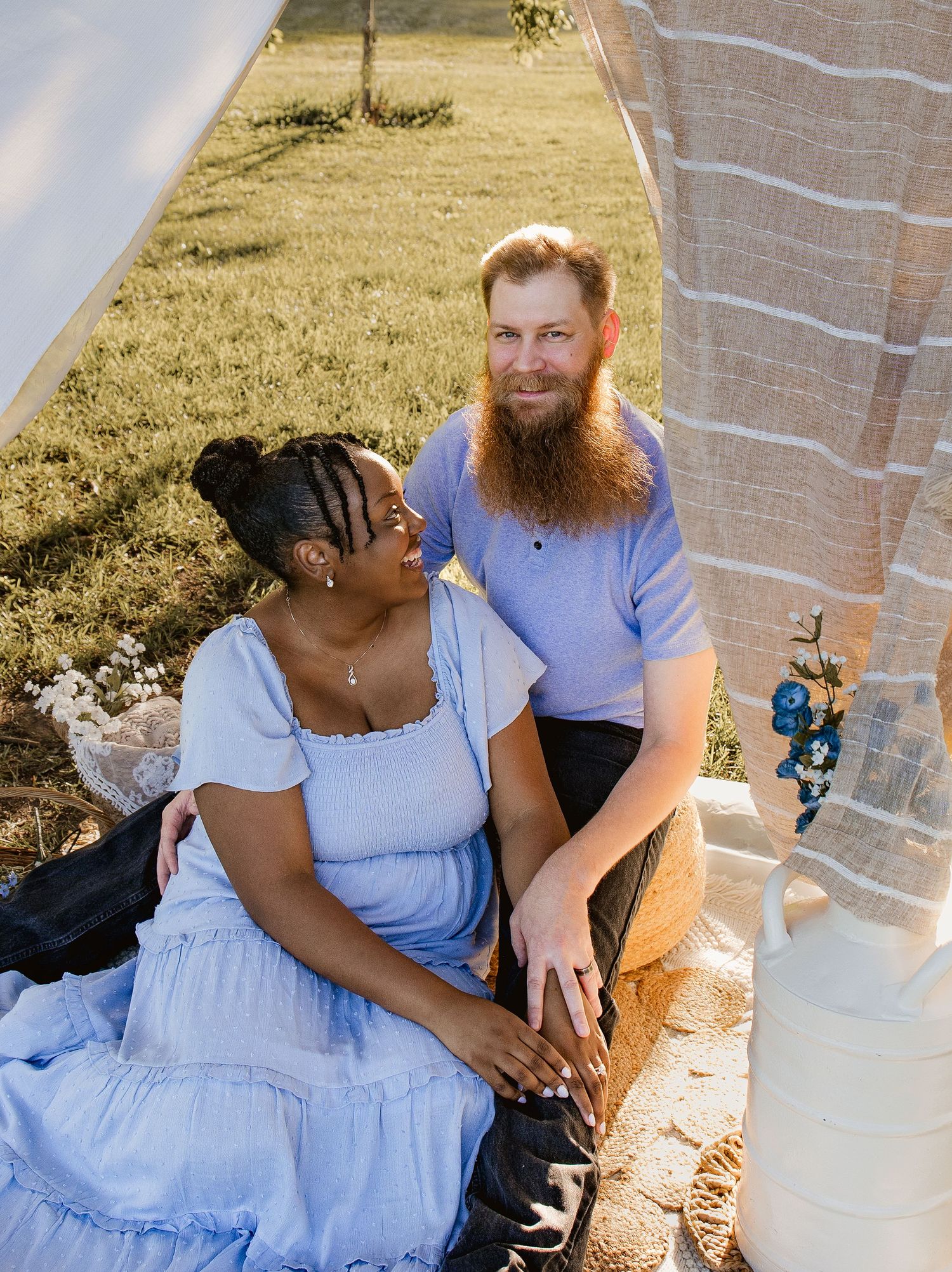 Couple sitting outdoors in a sunny field, woman in blue dress and bearded man smiling at each other.