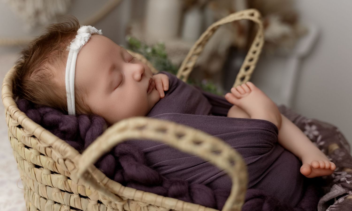 Peaceful newborn baby sleeping in a woven basket, swaddled in a purple wrap with a white headband.