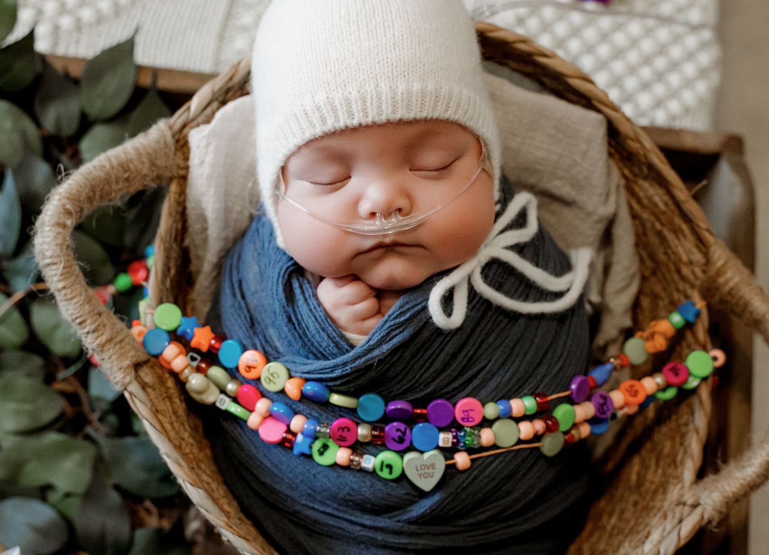 Sleeping newborn baby in white knit hat, wrapped in blue, nestled in wicker basket with colorful beaded necklace.