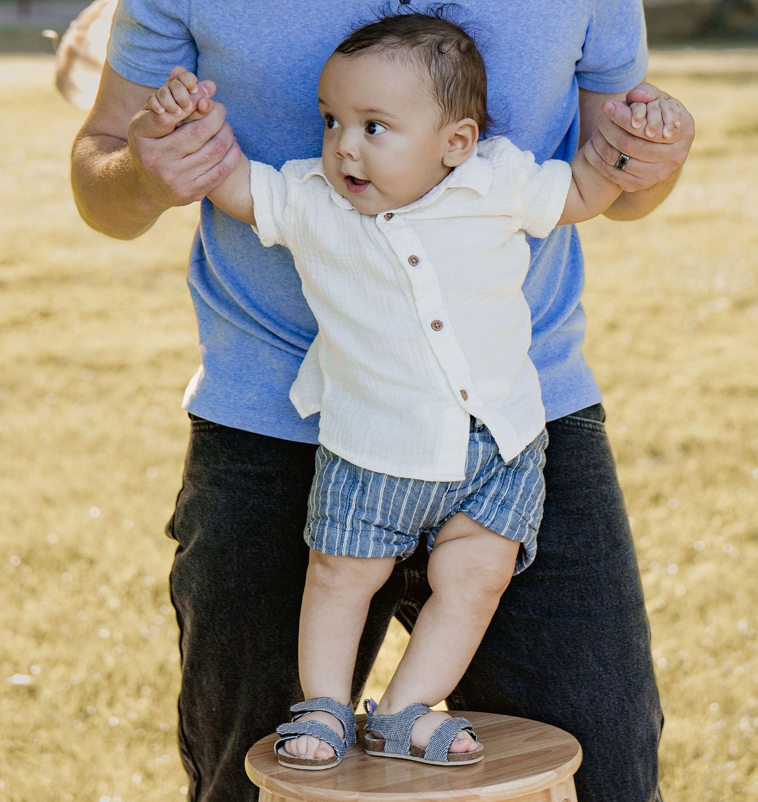 Baby boy in white shirt and blue shorts standing on a stool, supported by an adult in a blue shirt.