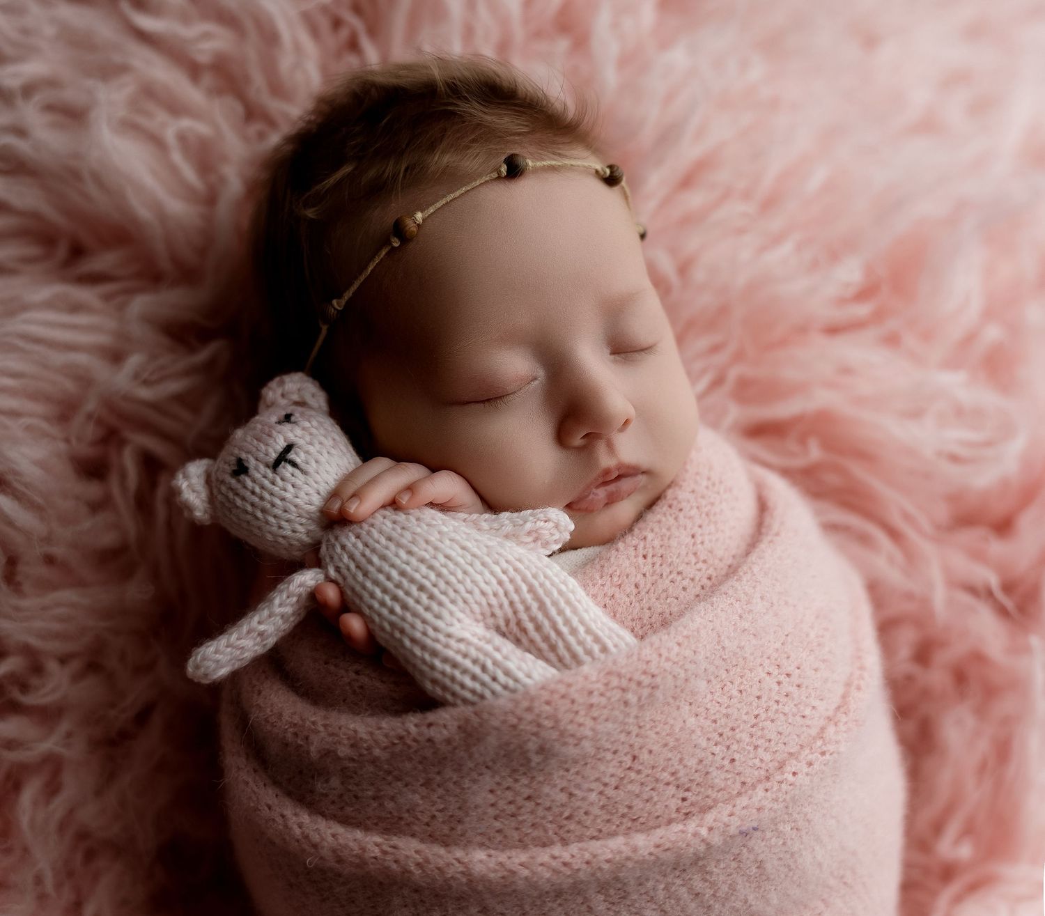 Newborn baby girl swaddled in pink wrap sleeping peacefully with knit bunny toy on fluffy pink fur backdrop.