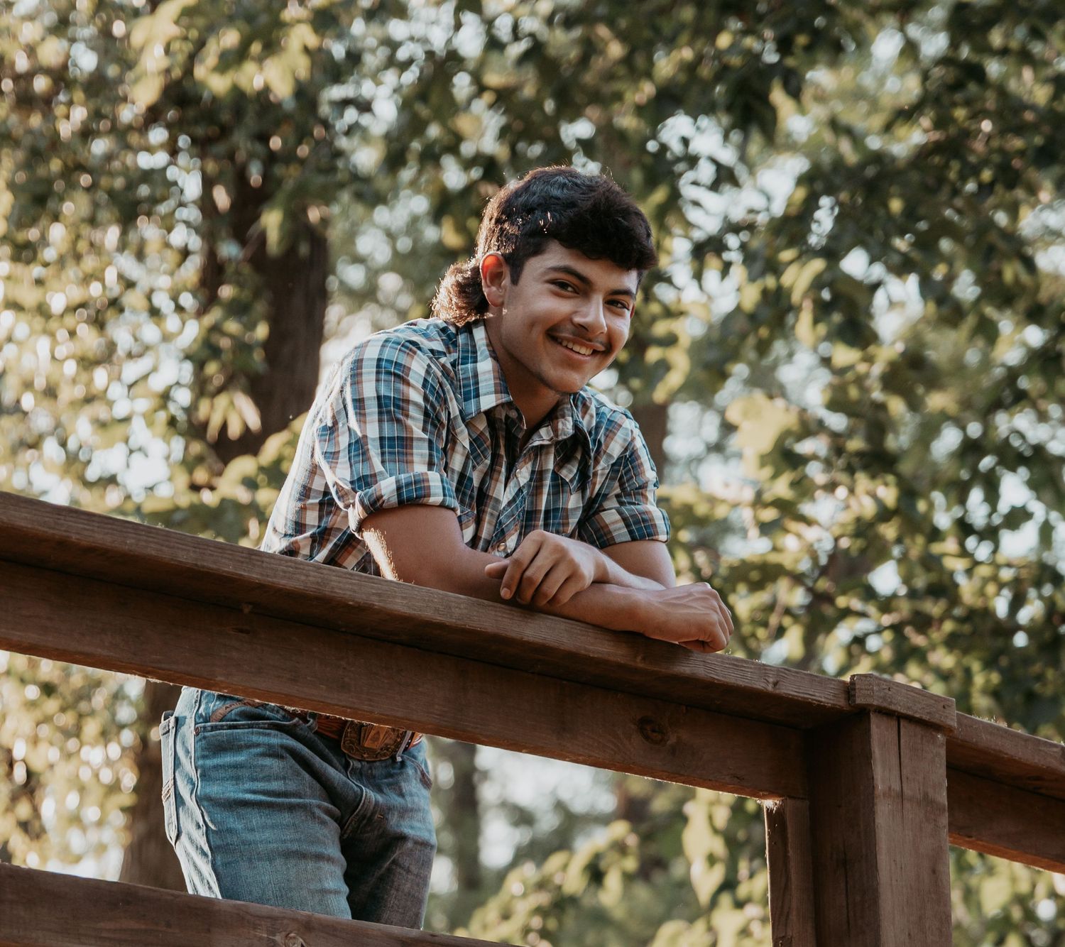 Smiling young man in plaid shirt leaning on wooden fence outdoors with green trees in background.