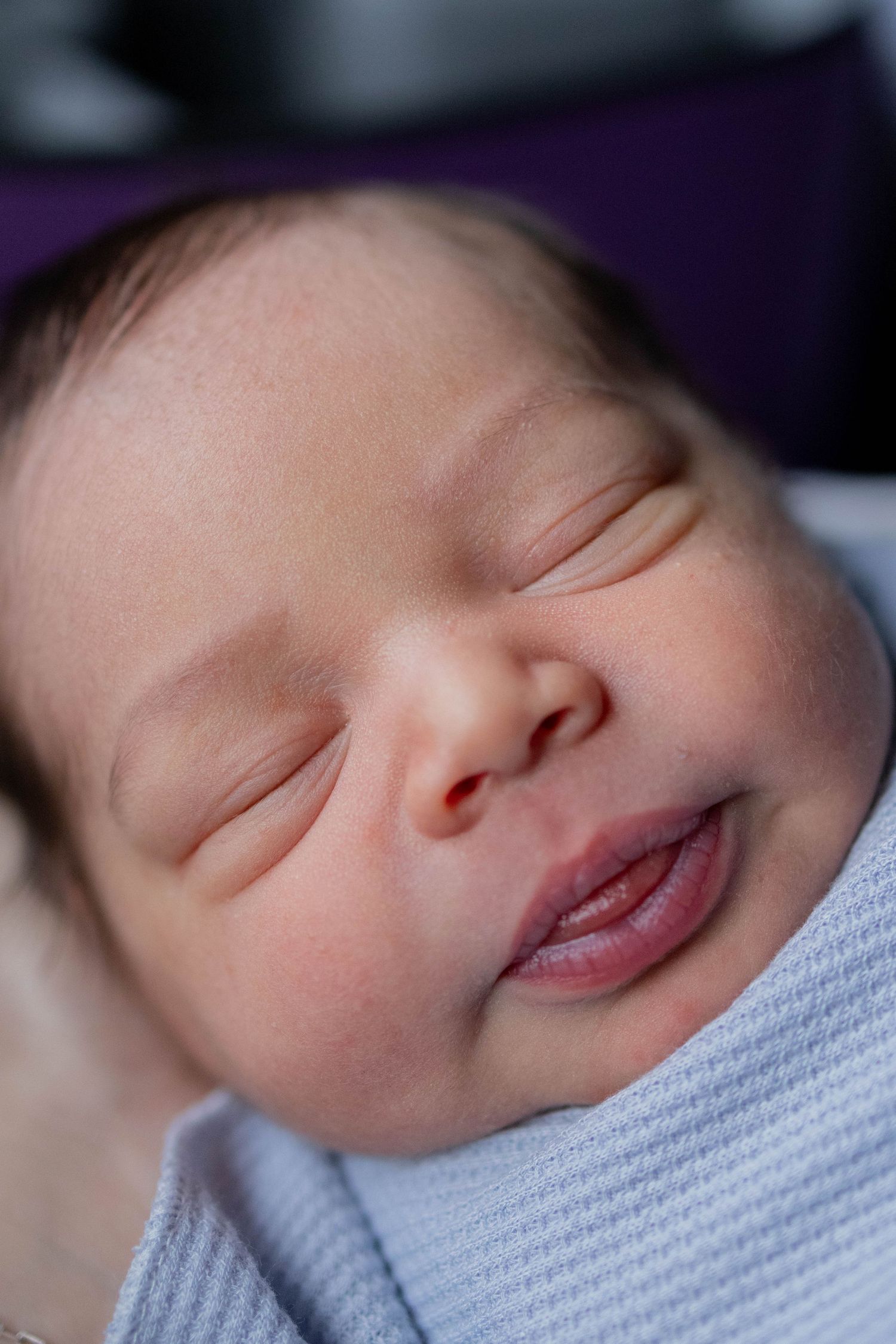 Peaceful sleeping newborn baby with dark hair, chubby cheeks, and full lips wrapped in a soft blue blanket.