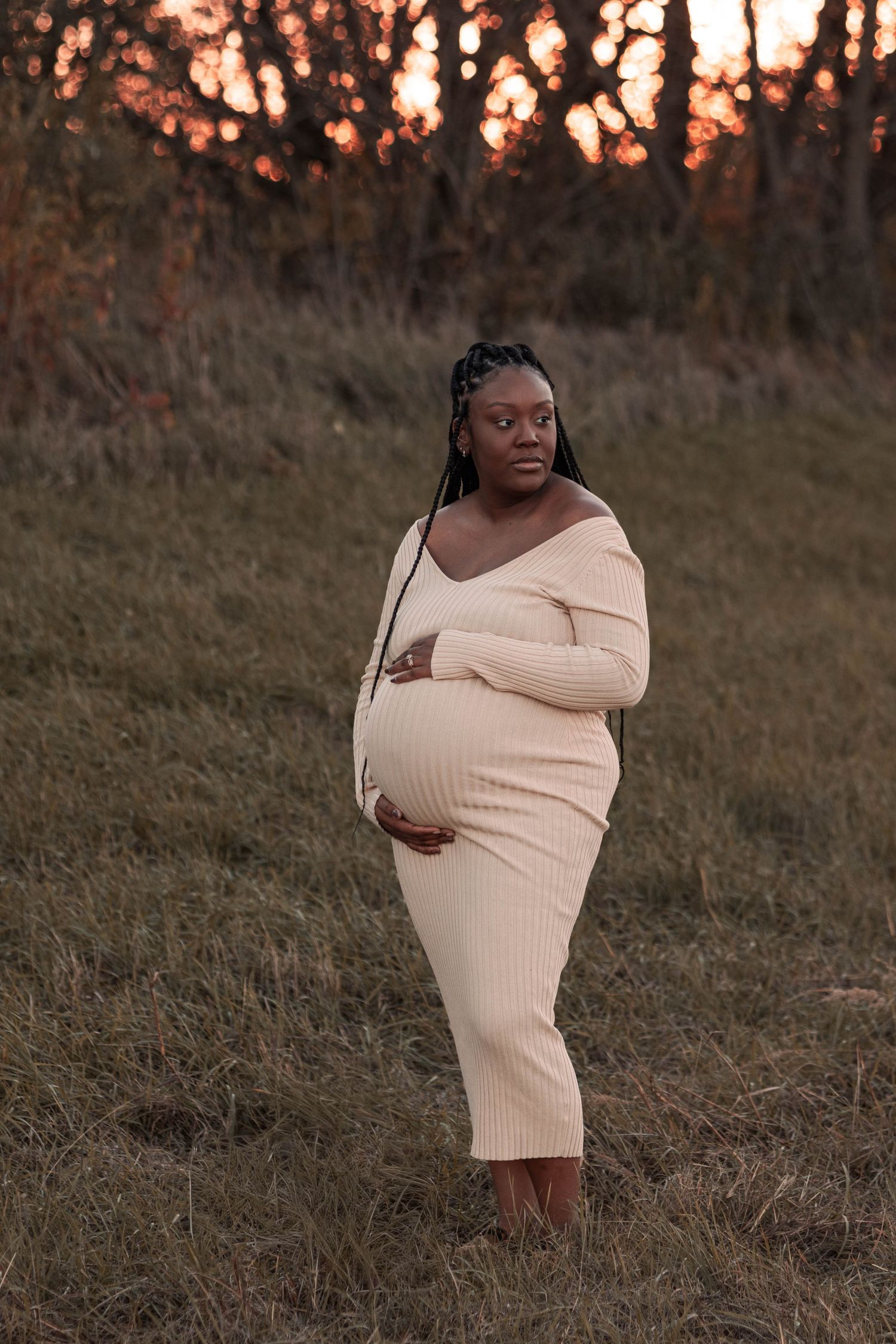 Pregnant Black woman in cream ribbed off-shoulder dress cradles belly outdoors at golden sunset.