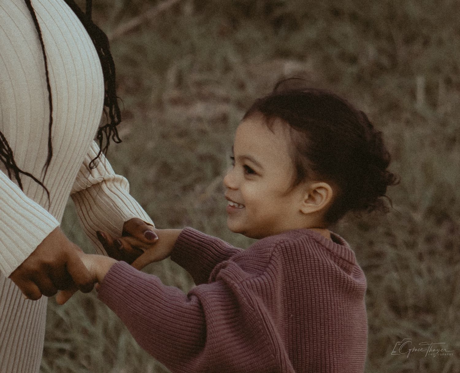 A smiling toddler in a mauve sweater holds their mothers hand outdoors in a field.