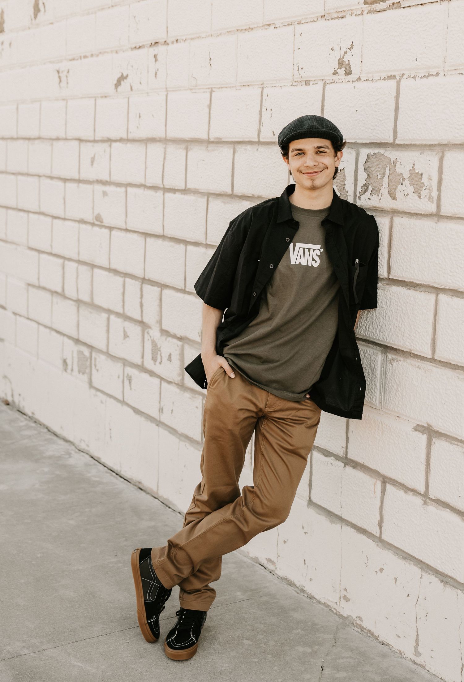 Young man in Vans tee, black shirt, tan pants, and beret leans against white brick wall smiling.