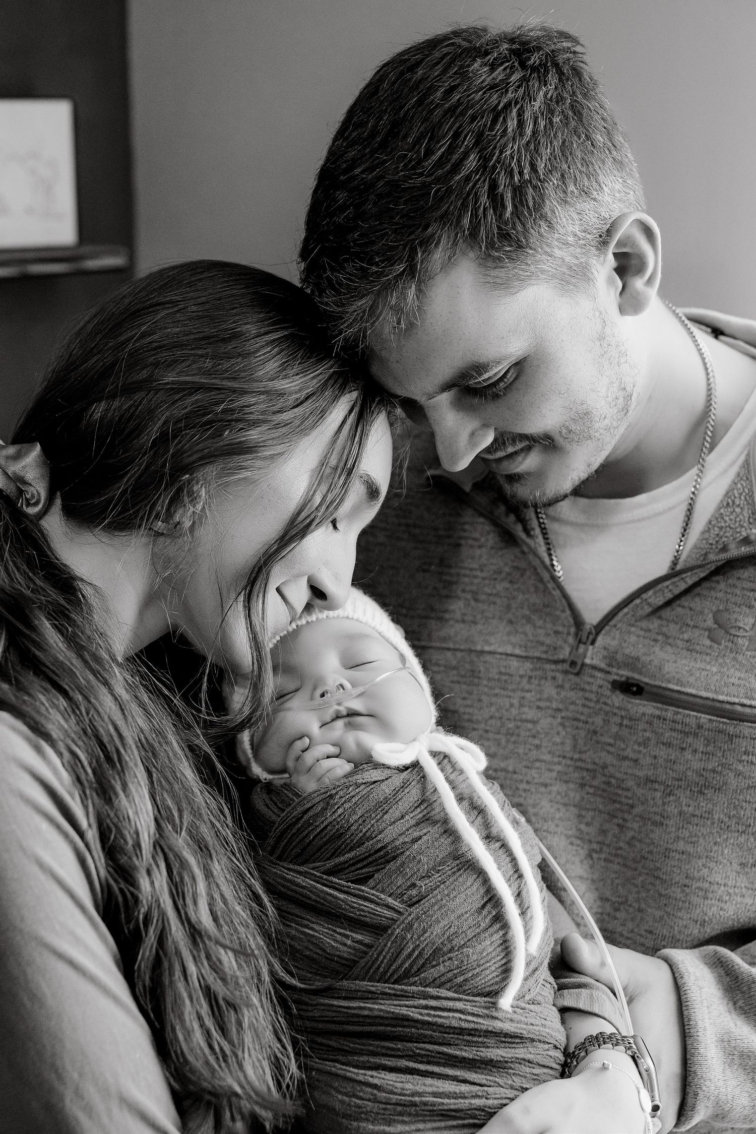 Black and white photo of new parents lovingly gazing at their sleeping newborn baby wrapped in a swaddle.