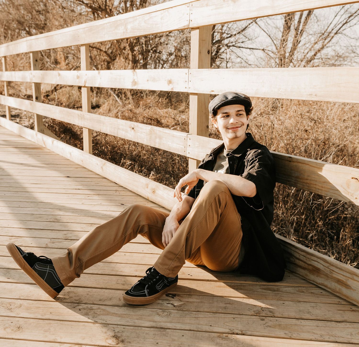 Young man in black shirt and tan pants sitting on a wooden bridge, smiling, wearing a flat cap and black sneakers.