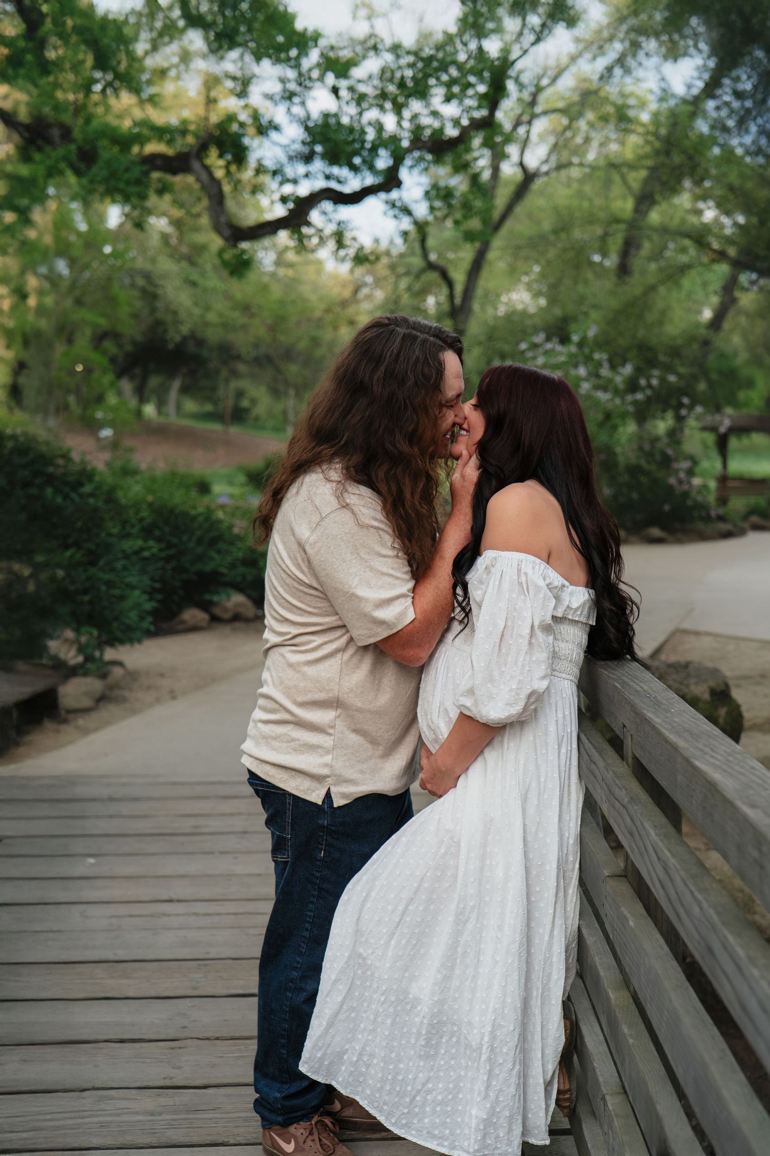 Romantic couple kissing on a wooden bridge in a lush park, woman in white off-shoulder maternity dress embracing long-haired partner.