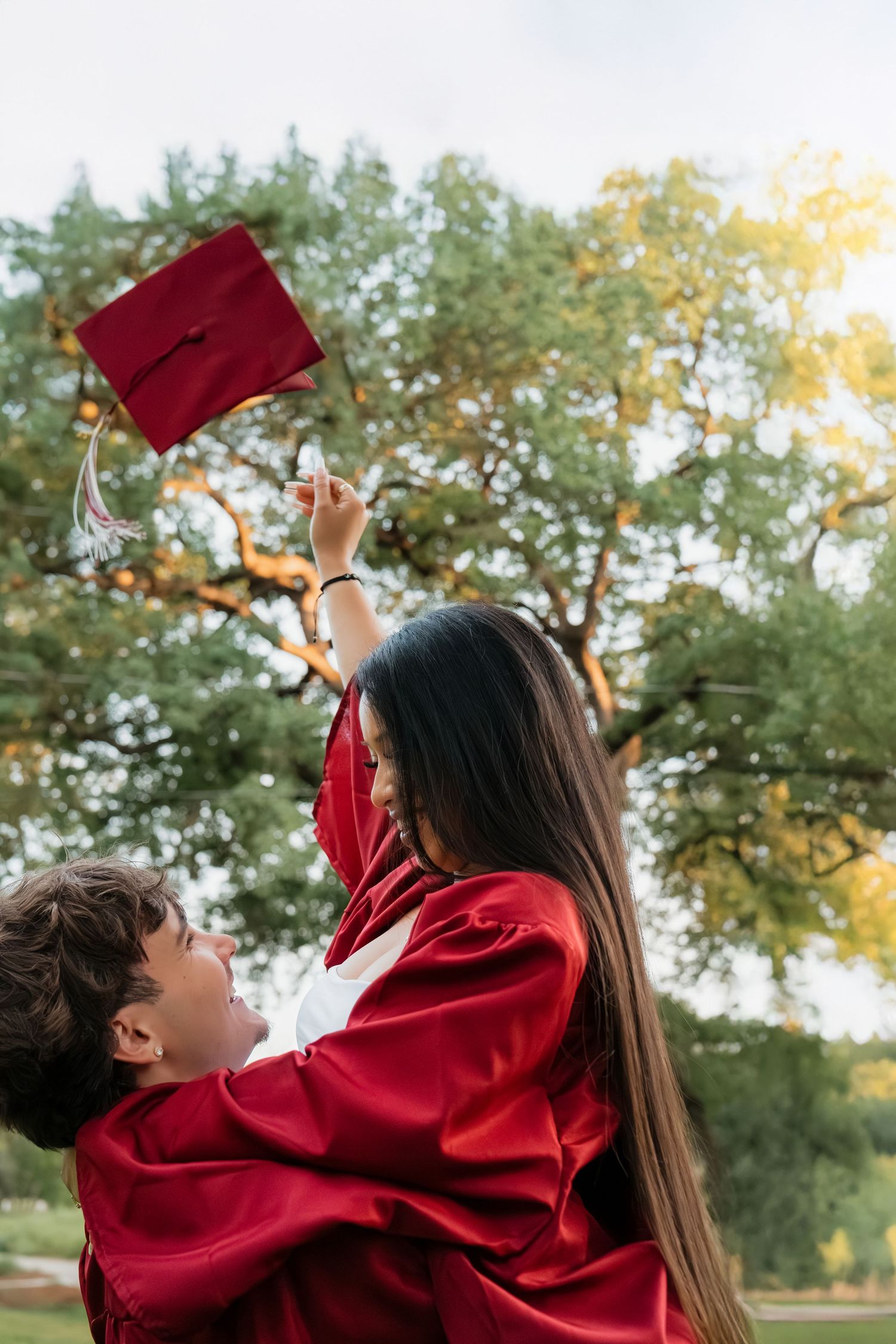 Two graduates in red gowns celebrate outdoors, one lifting a mortarboard cap into the air under sunny trees.