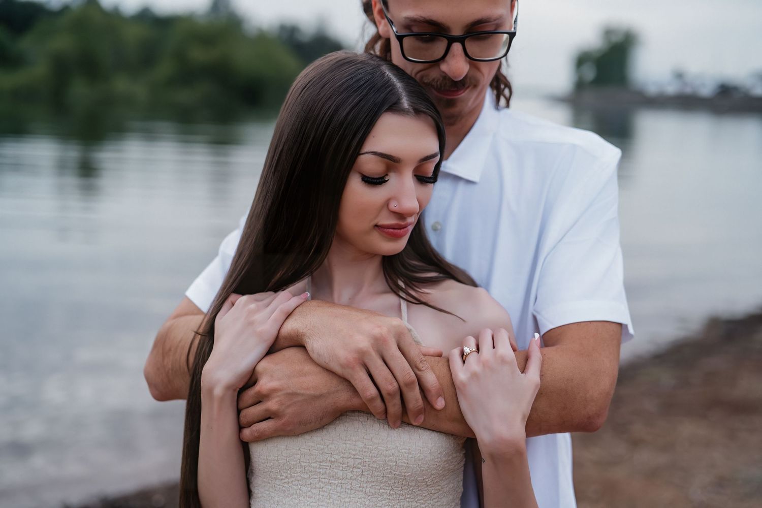 Romantic couple embracing near a lake, woman in strapless dress with man in white shirt holding her hands tenderly.