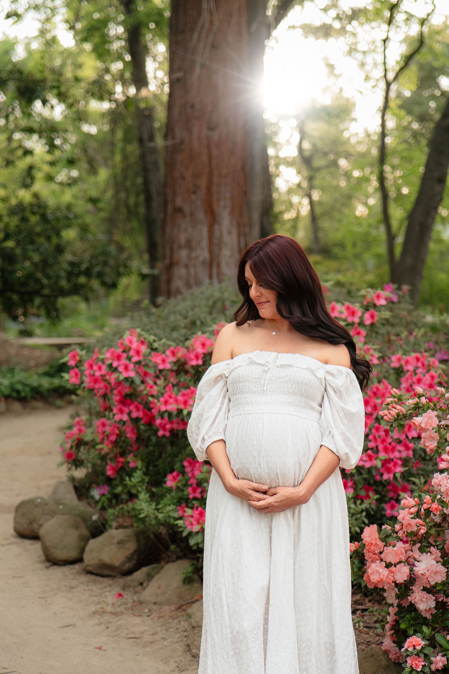 Pregnant woman in white off-shoulder dress cradles belly among pink flowers in sunlit garden.