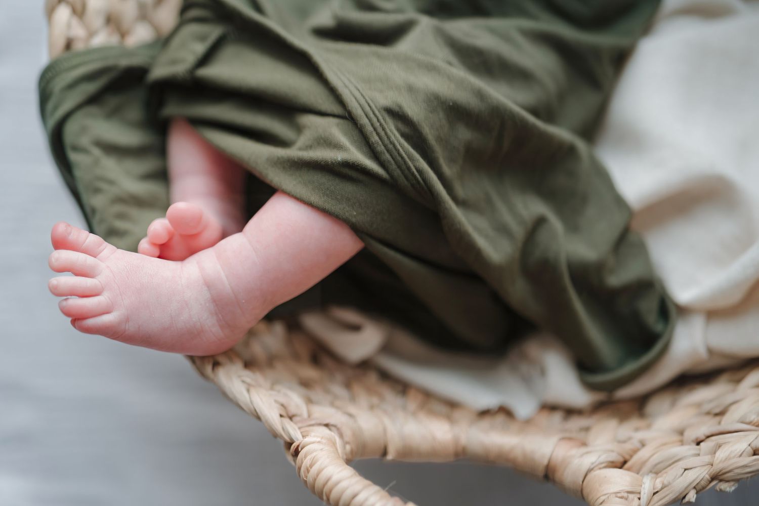 A tiny baby foot peeks out from under an olive green blanket while resting on a woven surface.