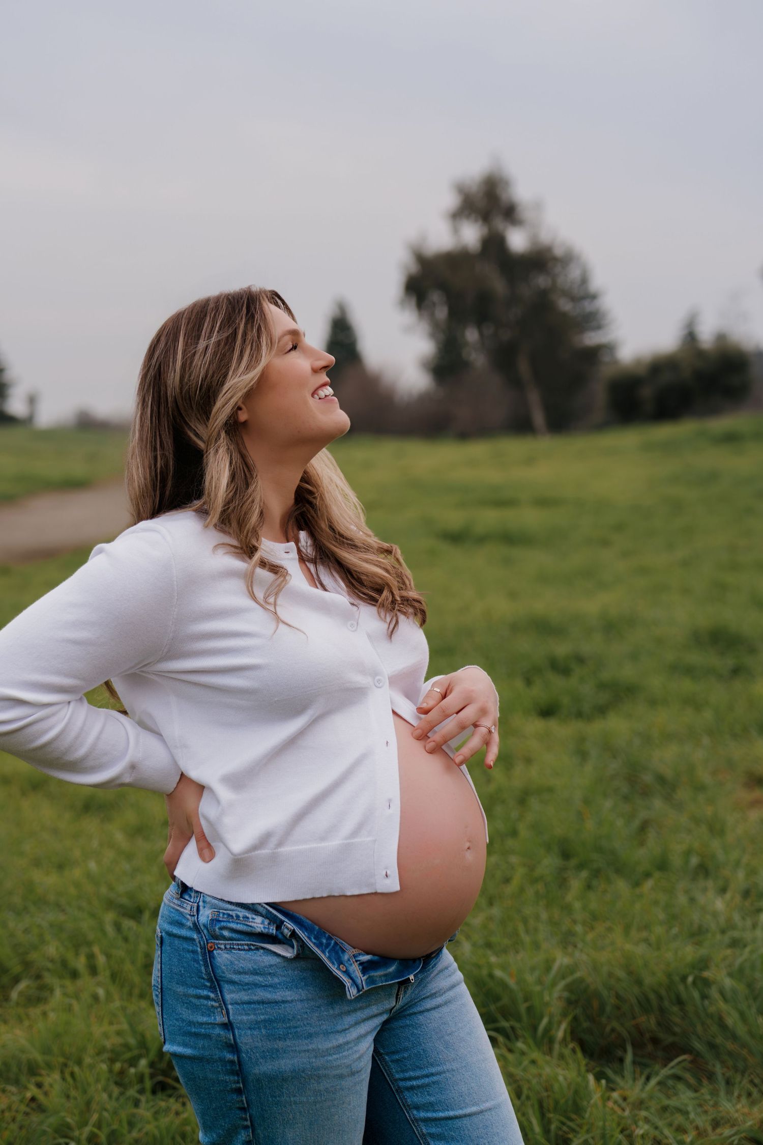 Smiling pregnant woman in white cardigan and jeans cradling her baby bump outdoors in a green field.