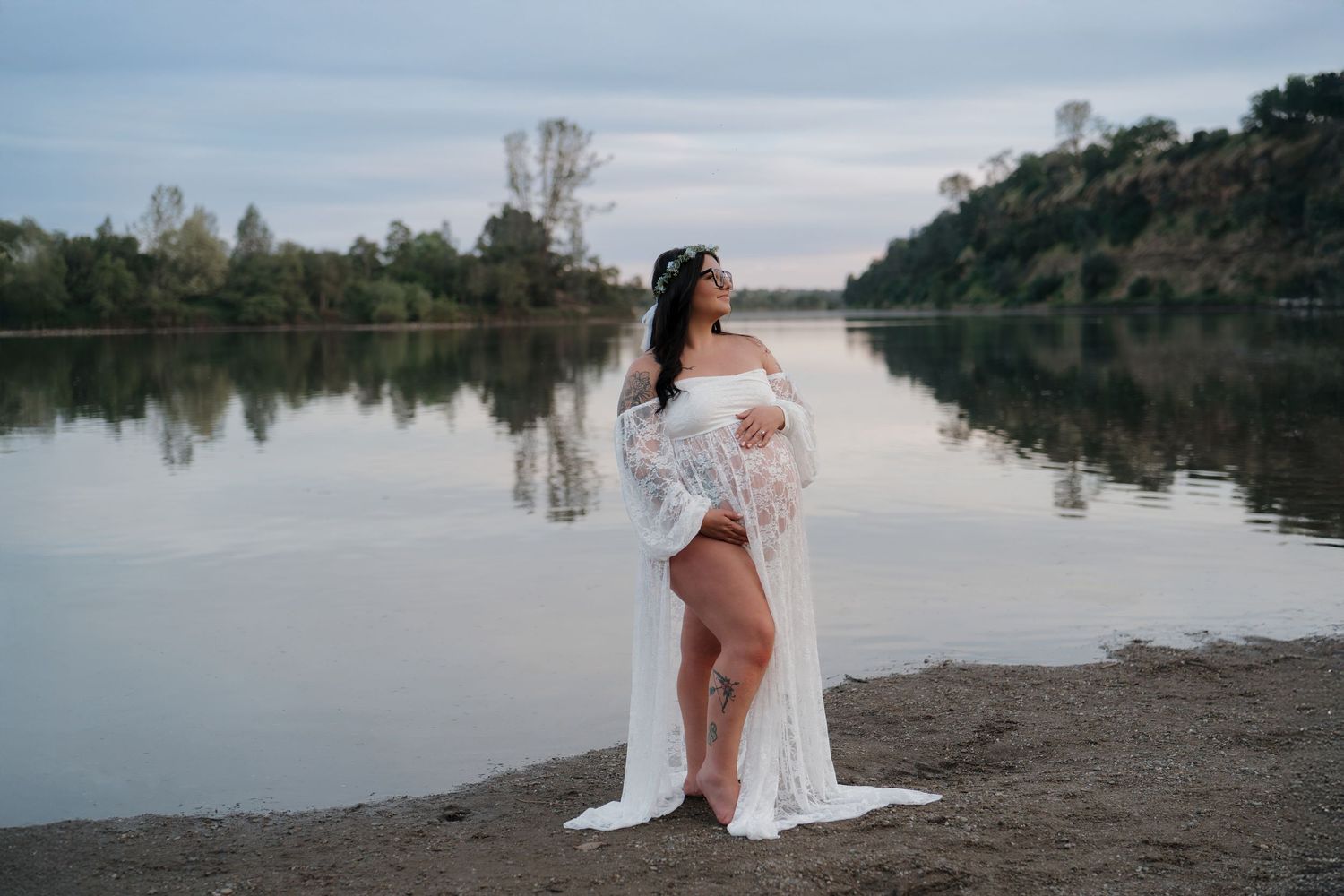 Pregnant woman in white lace gown poses gracefully by a serene river at dusk, showcasing a maternity photoshoot.